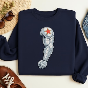 May include: Navy blue sweatshirt featuring a detailed illustration of a silver and gray robotic arm with a red star on the shoulder. The design is centered on the front of the long-sleeved garment.
