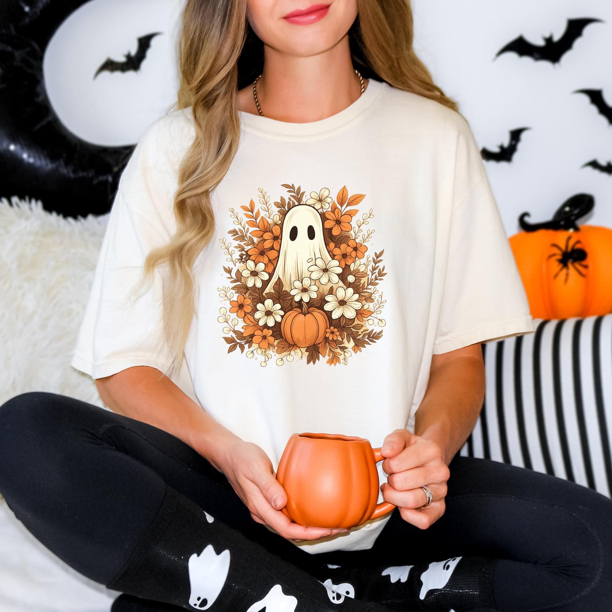A woman wearing a white t-shirt with a graphic of a ghost surrounded by flowers and a pumpkin, holding a mug in her hands.