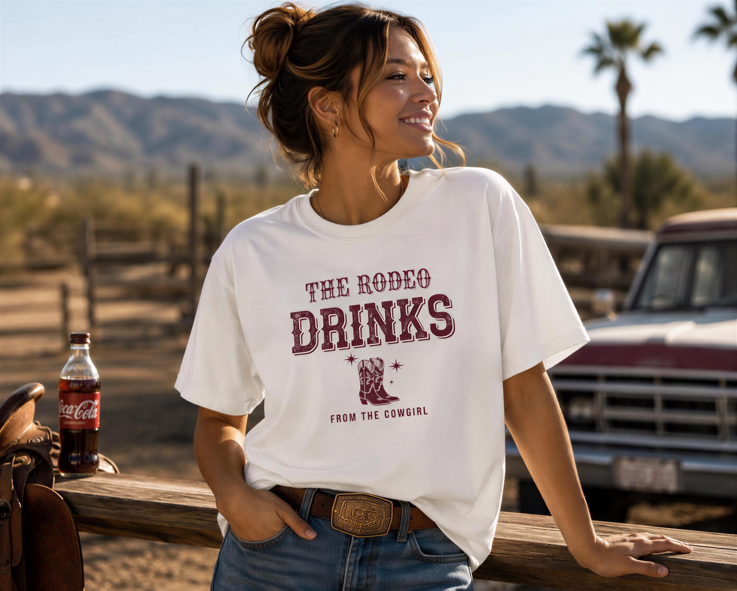 A woman wearing a white t-shirt with the text "THE RODEO DRINKS FROM THE COWGIRL" is leaning against a wooden fence, smiling and looking to the side.