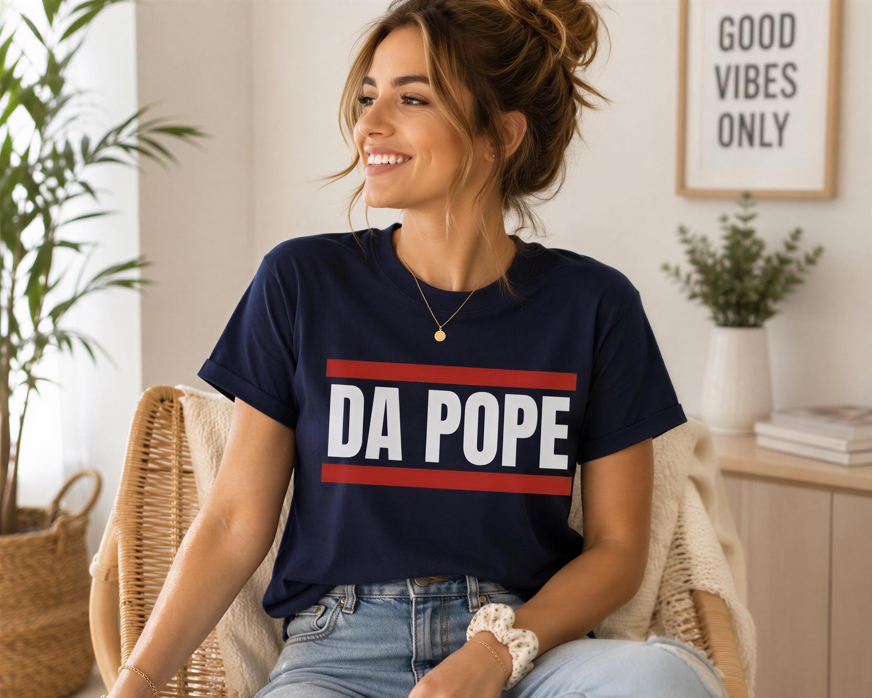 A woman wearing a navy blue t-shirt with the text "DA POPE" is sitting in a chair, smiling and looking to the side.