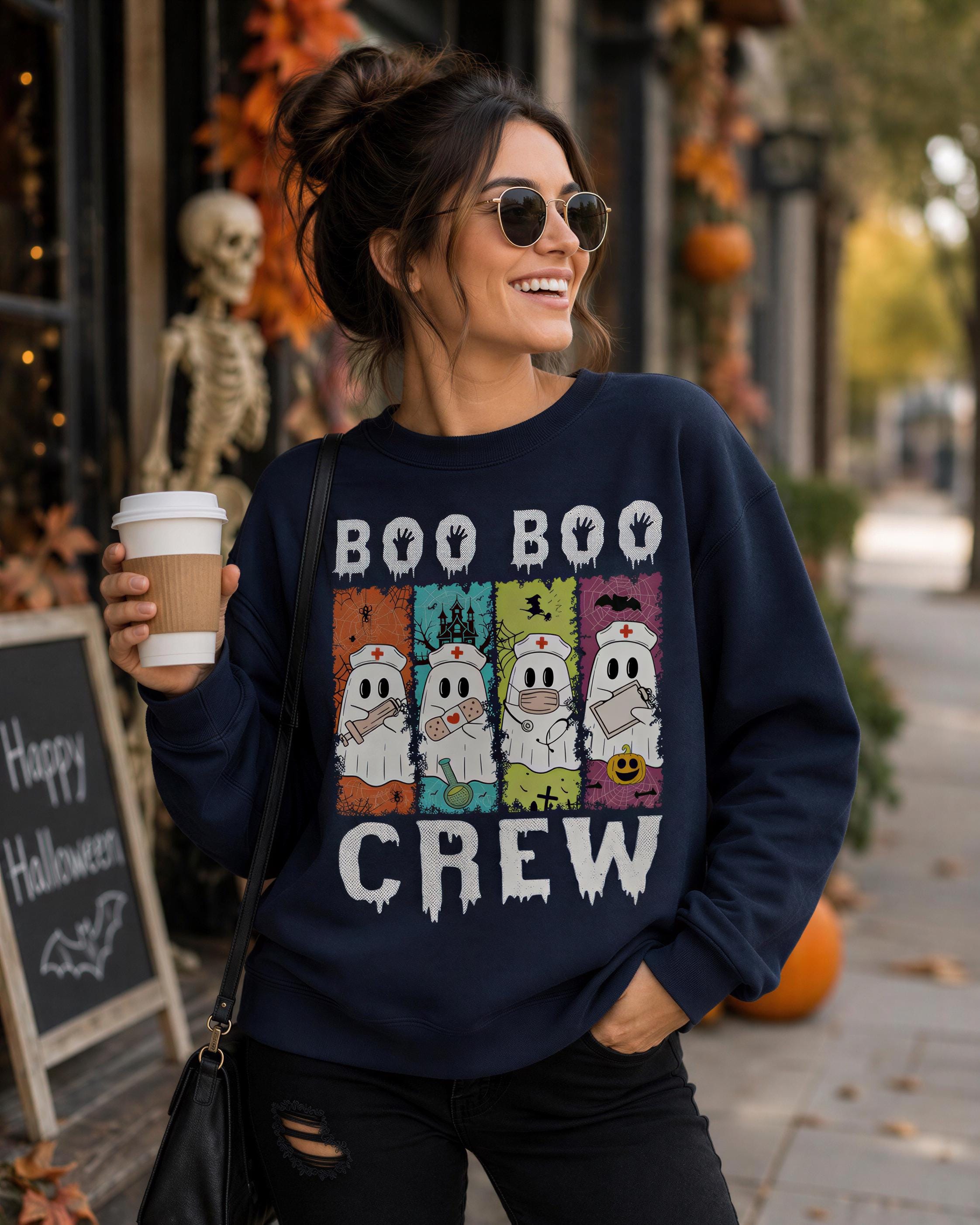 A woman wearing a navy blue sweater with a graphic of a ghost crew and the text "BOO BOO CREW" is standing on a sidewalk, holding a coffee cup and smiling.