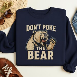 May include: Navy blue sweatshirt with the text "DON'T POKE THE BEAR" above a graphic of a roaring bear. The bear is light brown with a dark brown outline. The text is in a bold, sans-serif font.