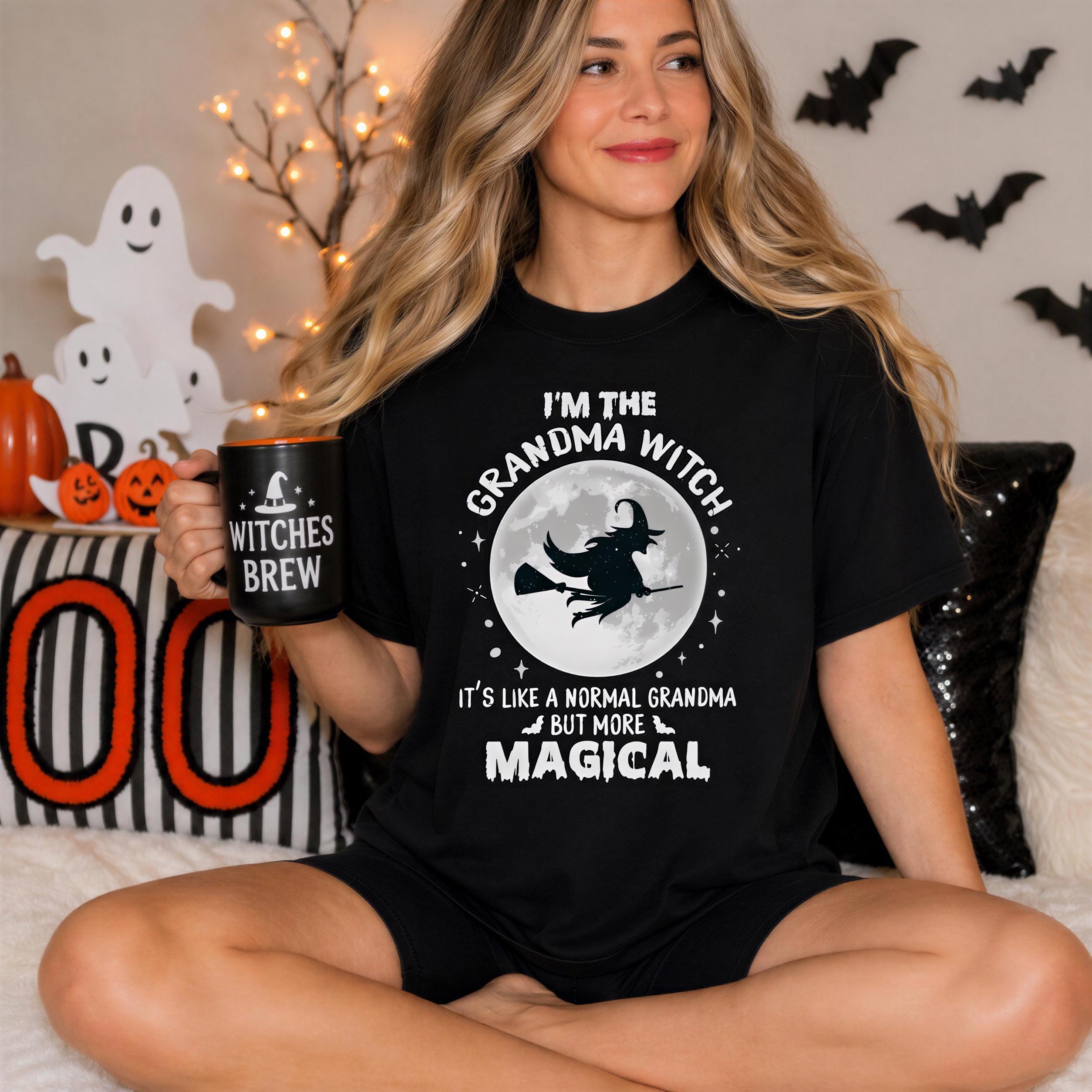 A woman wearing a black t-shirt with a Halloween-themed design is sitting on a couch, holding a mug with the text "I'm the GRANDMA WITCH" and "IT'S LIKE A NORMAL GRANDMA BUT MORE MAGICAL".