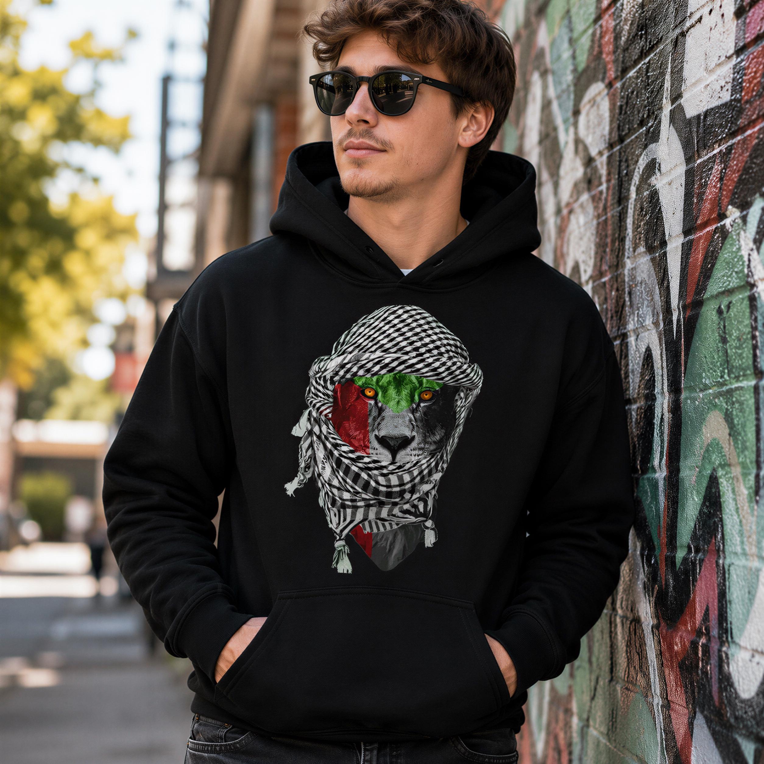 A man wearing a black hoodie with a graphic of a cat wearing a scarf and sunglasses stands in front of a colorful graffiti-covered wall.