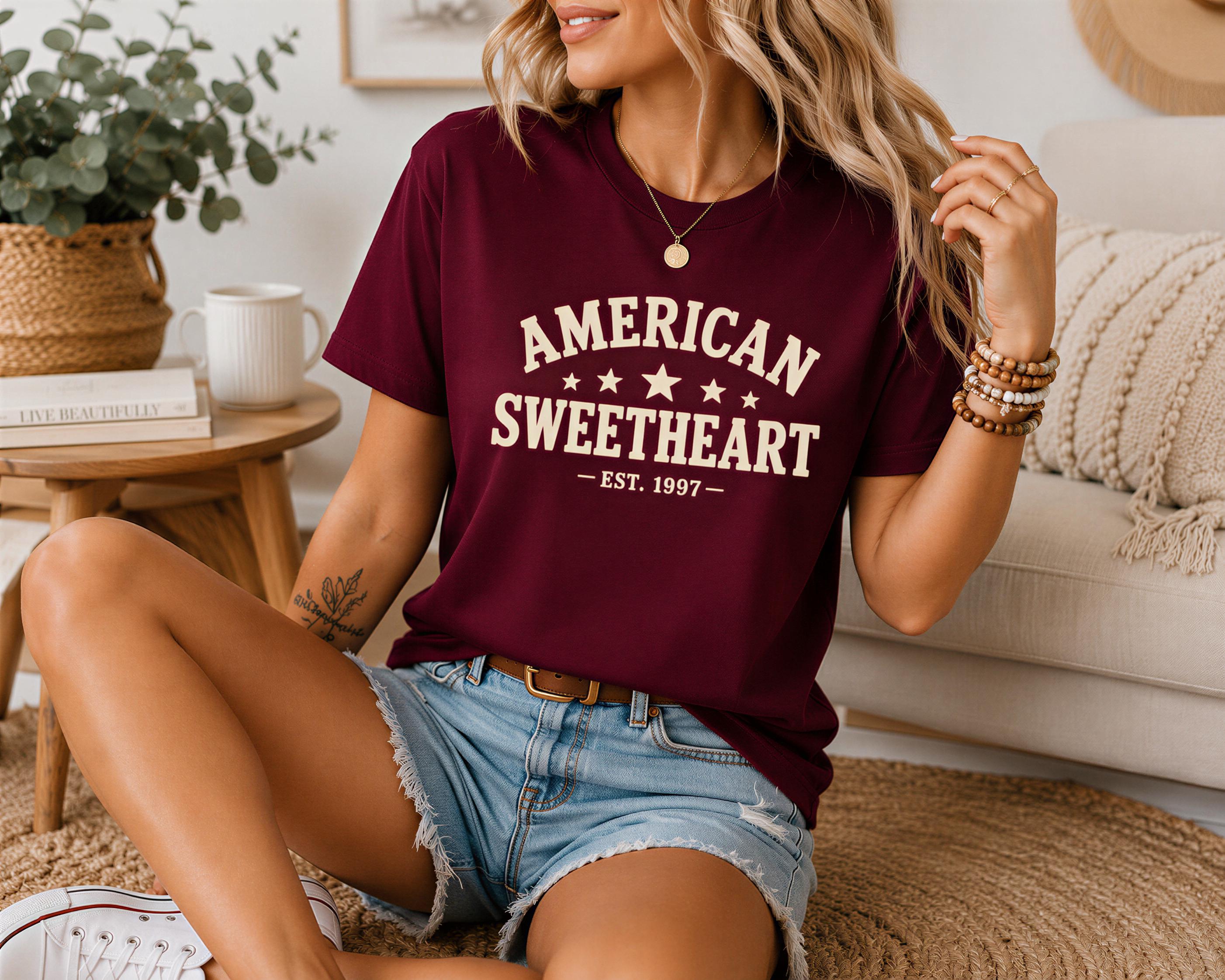 A woman wearing a maroon t-shirt with the text "AMERICAN SWEETHEART" and denim shorts is sitting on the floor in a living room.