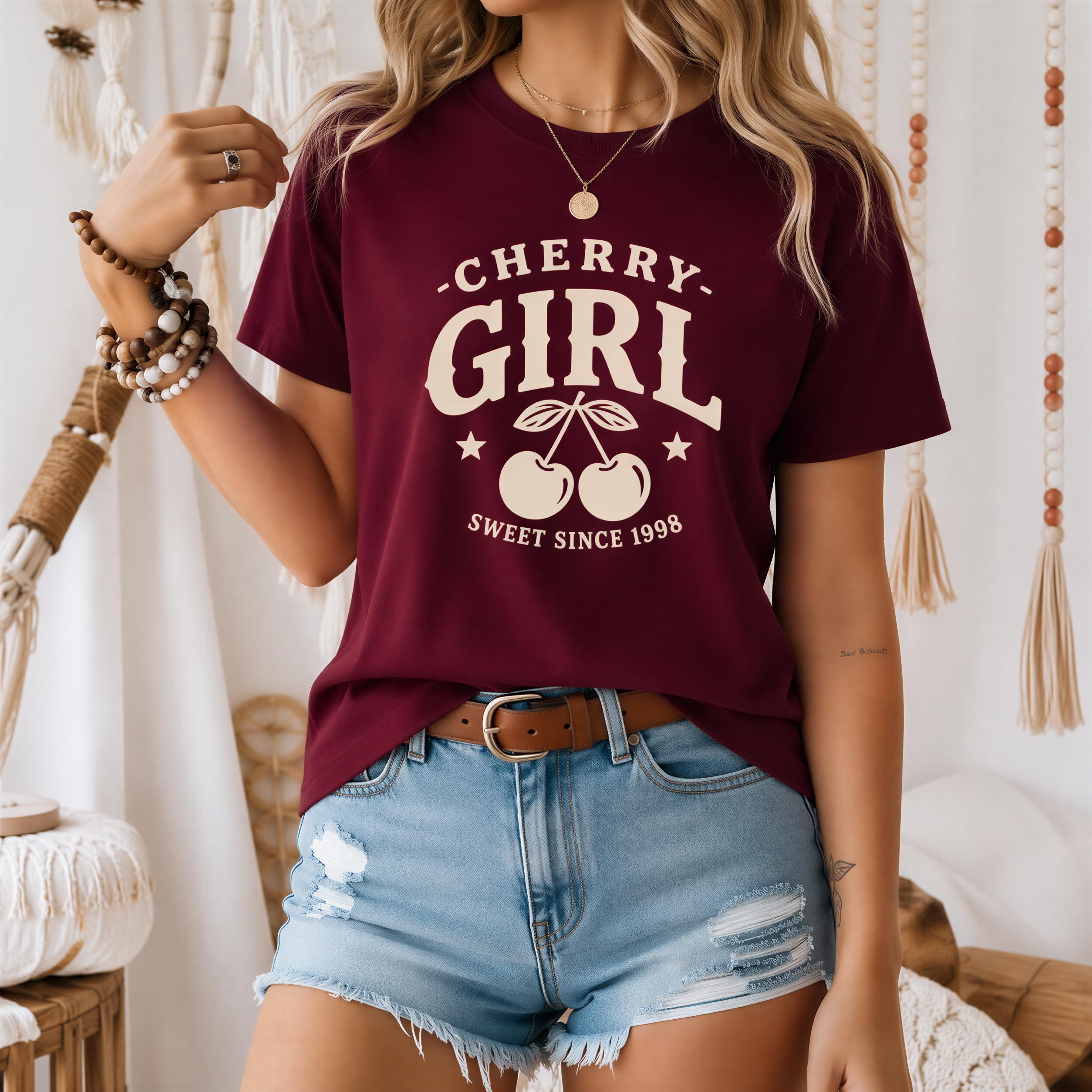 A woman wearing a burgundy t-shirt with the text "CHERRY GIRL" and denim shorts, standing in front of a beaded curtain.
