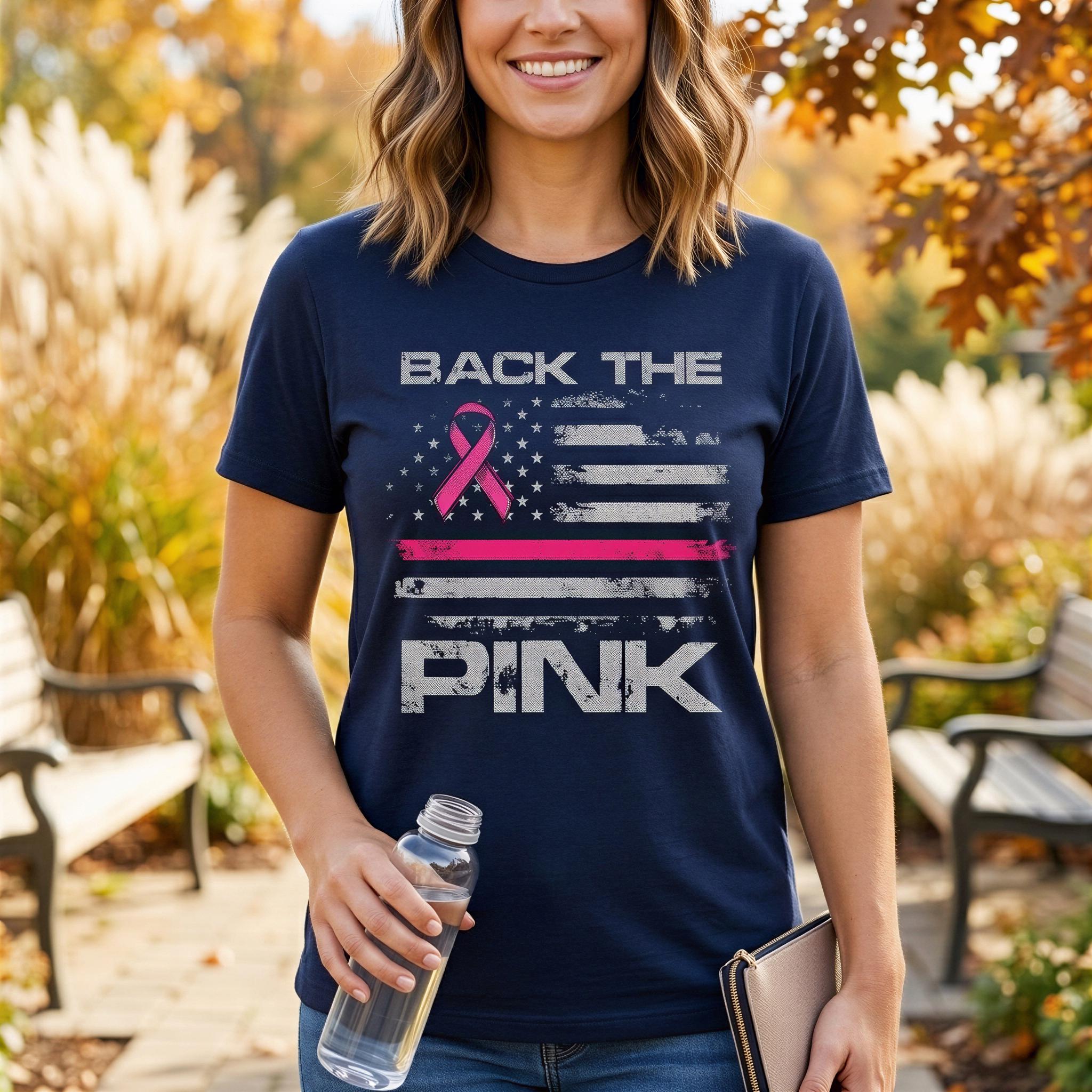A woman wearing a navy blue t-shirt with the text "BACK THE PINK" and a pink ribbon graphic, standing in a park with autumn foliage in the background.