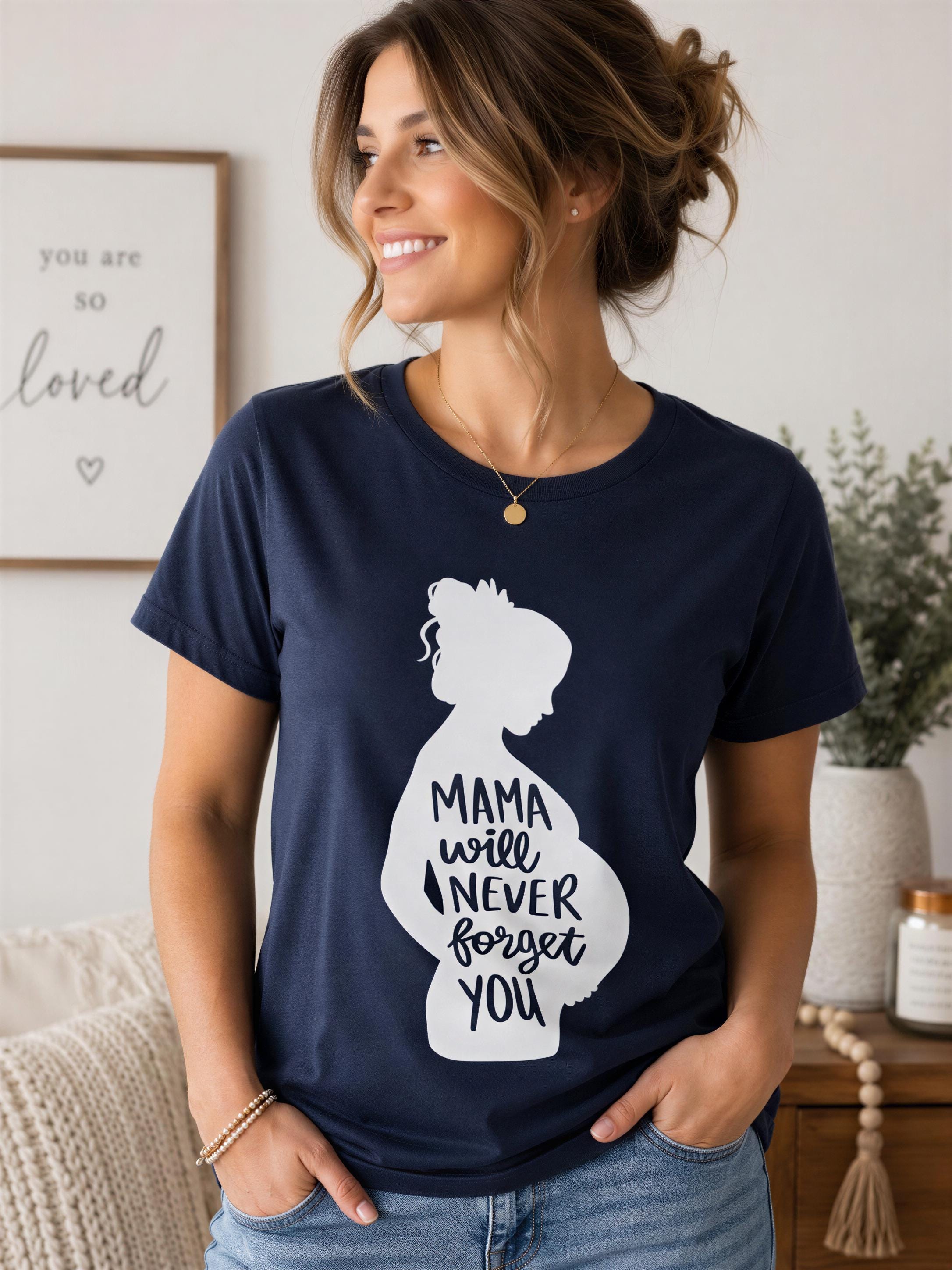 A woman wearing a navy blue t-shirt with a graphic design of a silhouette of a pregnant woman and the text "Mama will never forget you" is smiling and looking to the side.