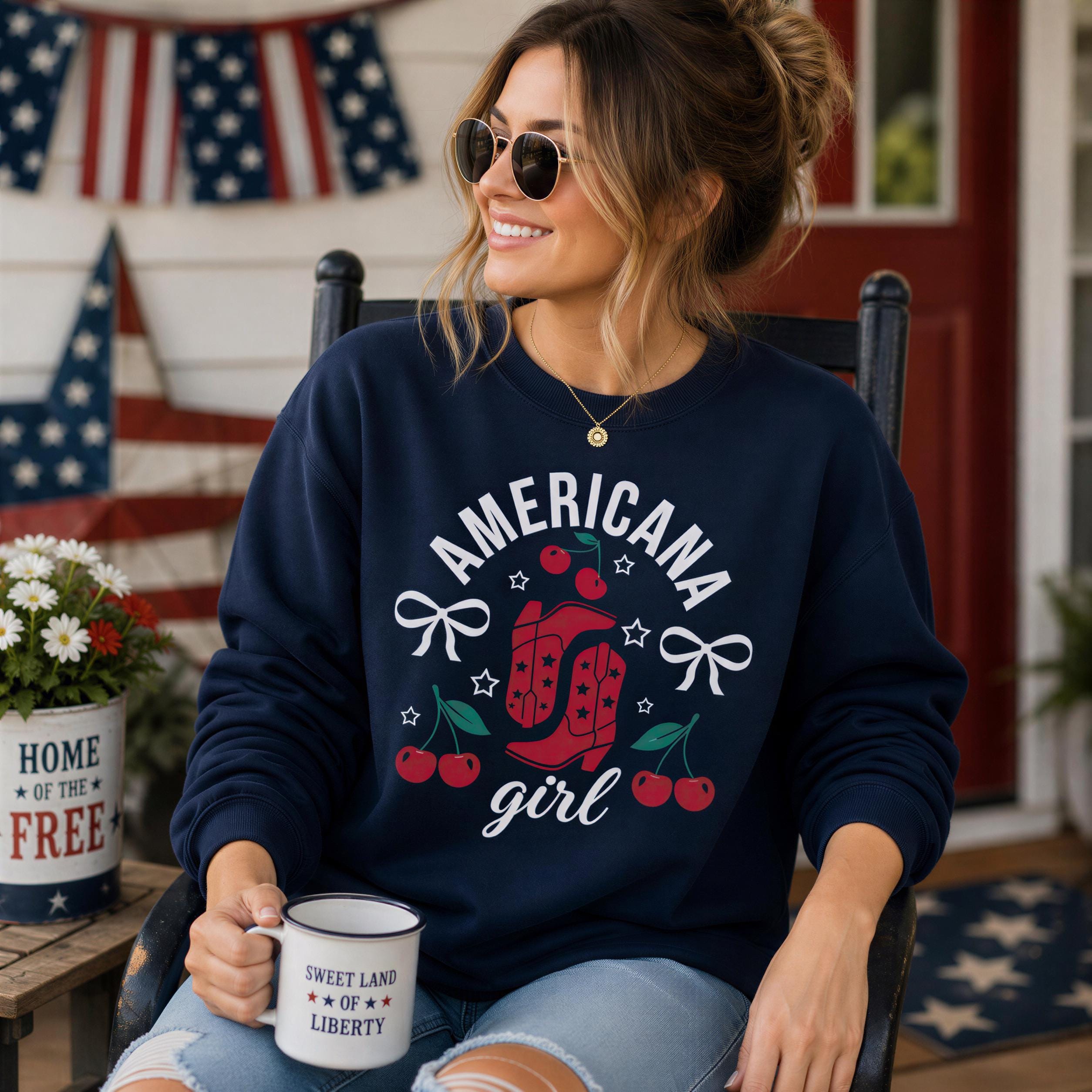 A woman wearing a navy blue sweatshirt with the text "AMERICANA GIRL" and a graphic of a red cowboy boot, sunglasses, and cherries, sitting on a porch with a mug and flowers in the background.