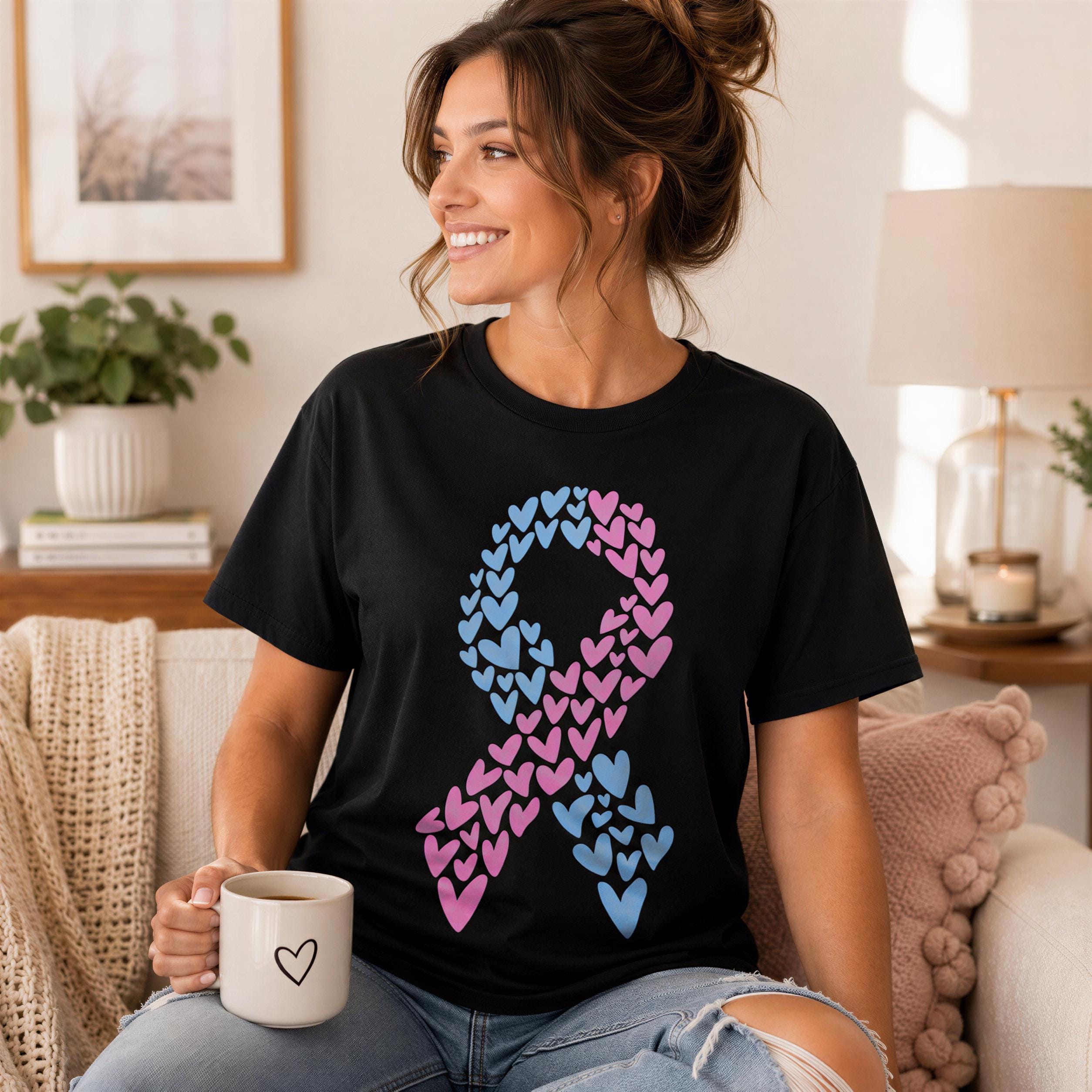 A woman sitting on a couch, smiling and holding a coffee mug, wearing a black t-shirt with a pink and blue heart ribbon design.