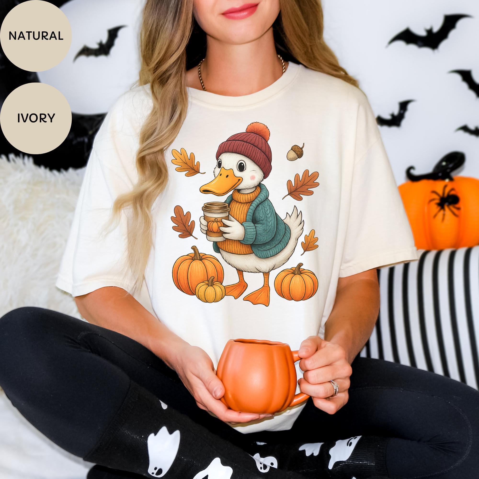 A woman wearing a white t-shirt with a cartoon duck holding a coffee mug and surrounded by autumn leaves and pumpkins.