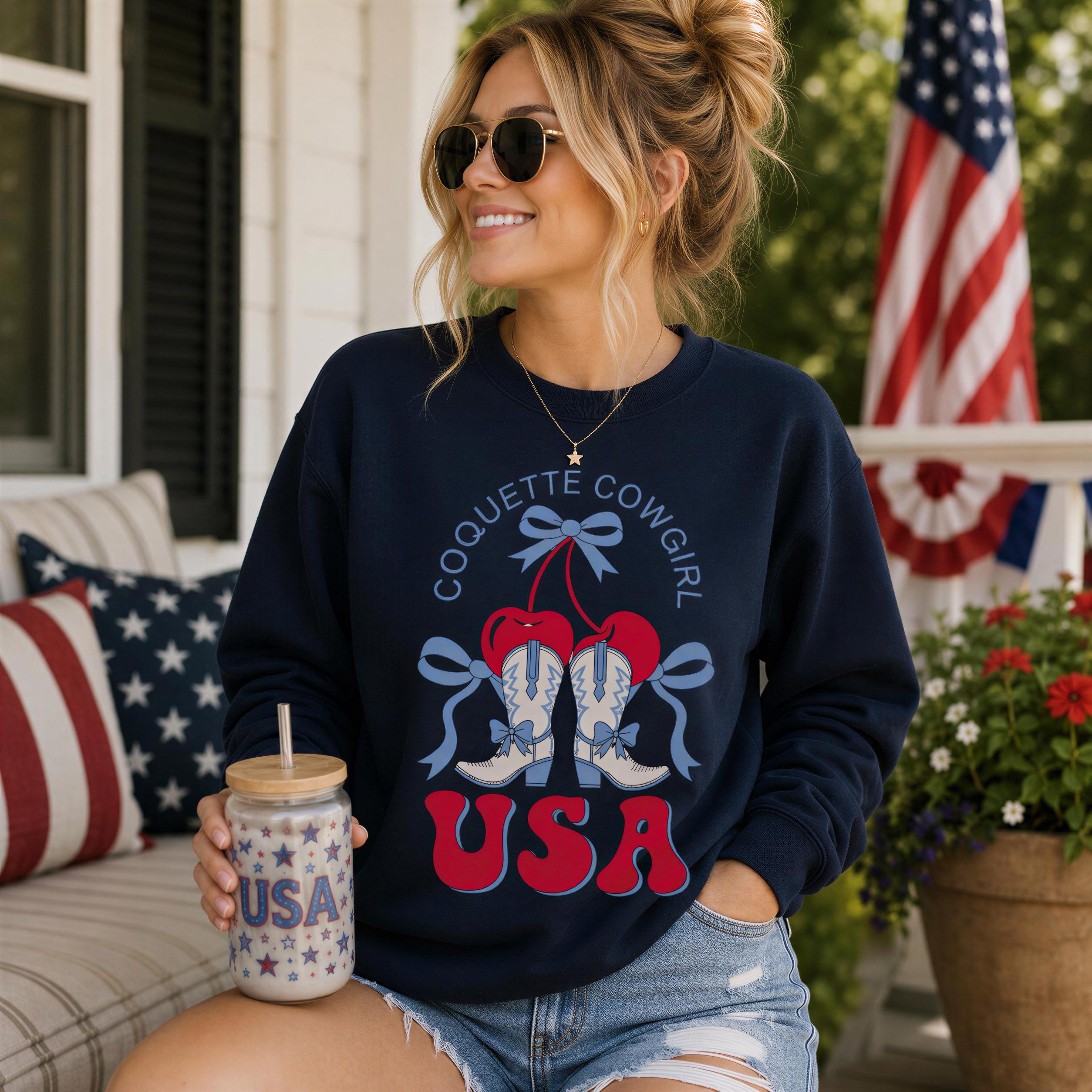 A woman wearing a navy blue sweatshirt with a graphic of cowboy boots and the text "Coquette Cowgirl USA" is sitting on a porch, holding a drink with a straw.