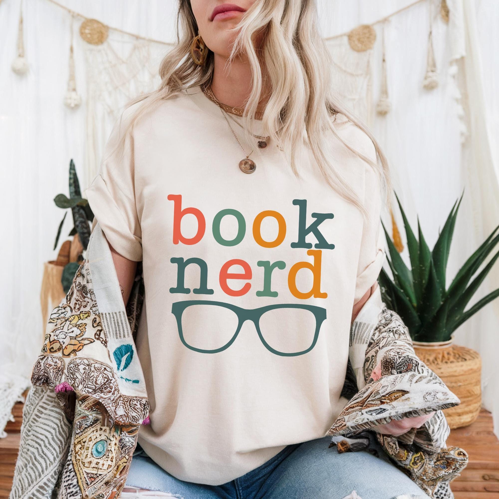 A woman wearing a white t-shirt with the text "book nerd" printed on it, sitting in front of a white curtain and surrounded by plants.