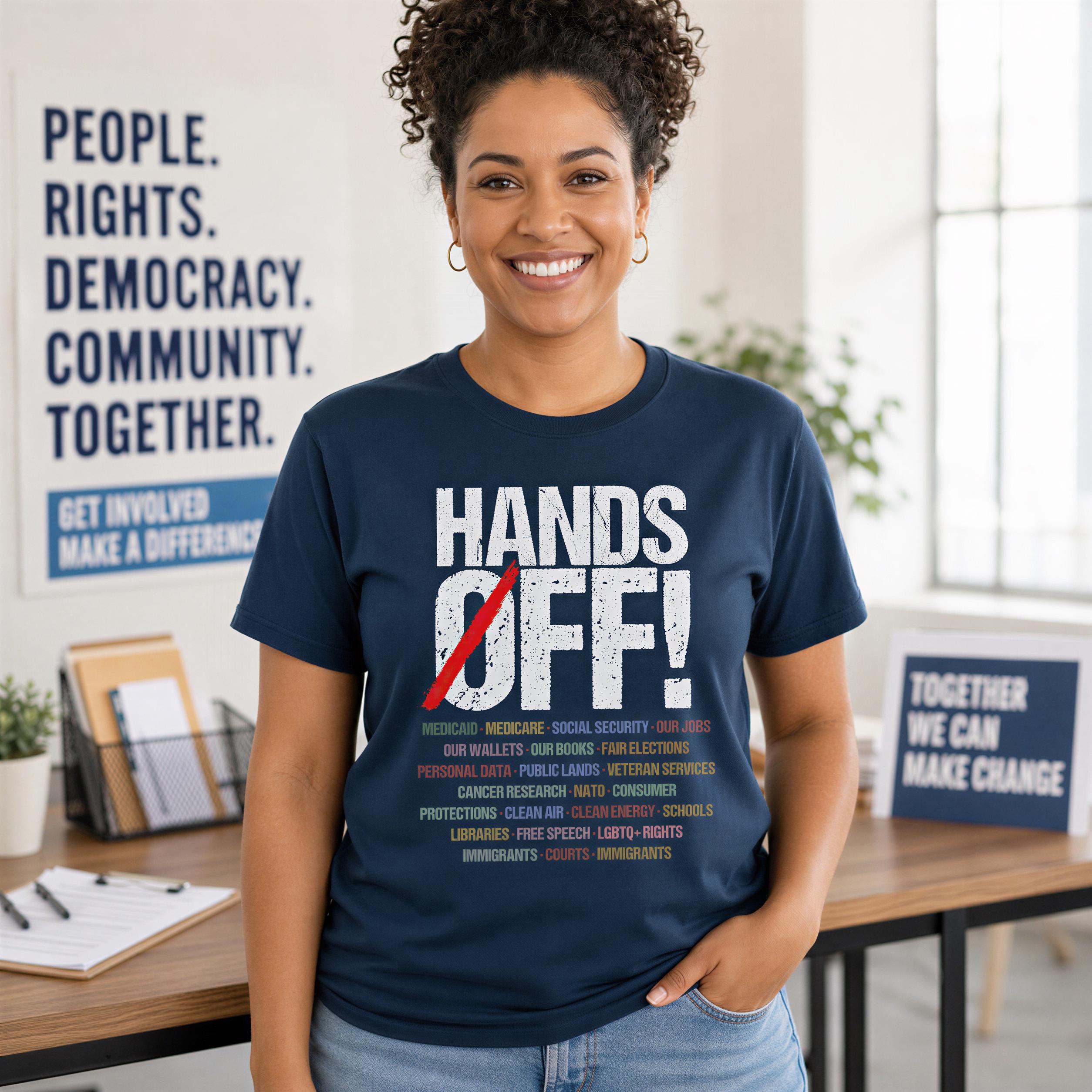 A smiling woman wearing a navy blue t-shirt with a bold statement printed on it, standing in an office setting with a desk and a plant in the background.