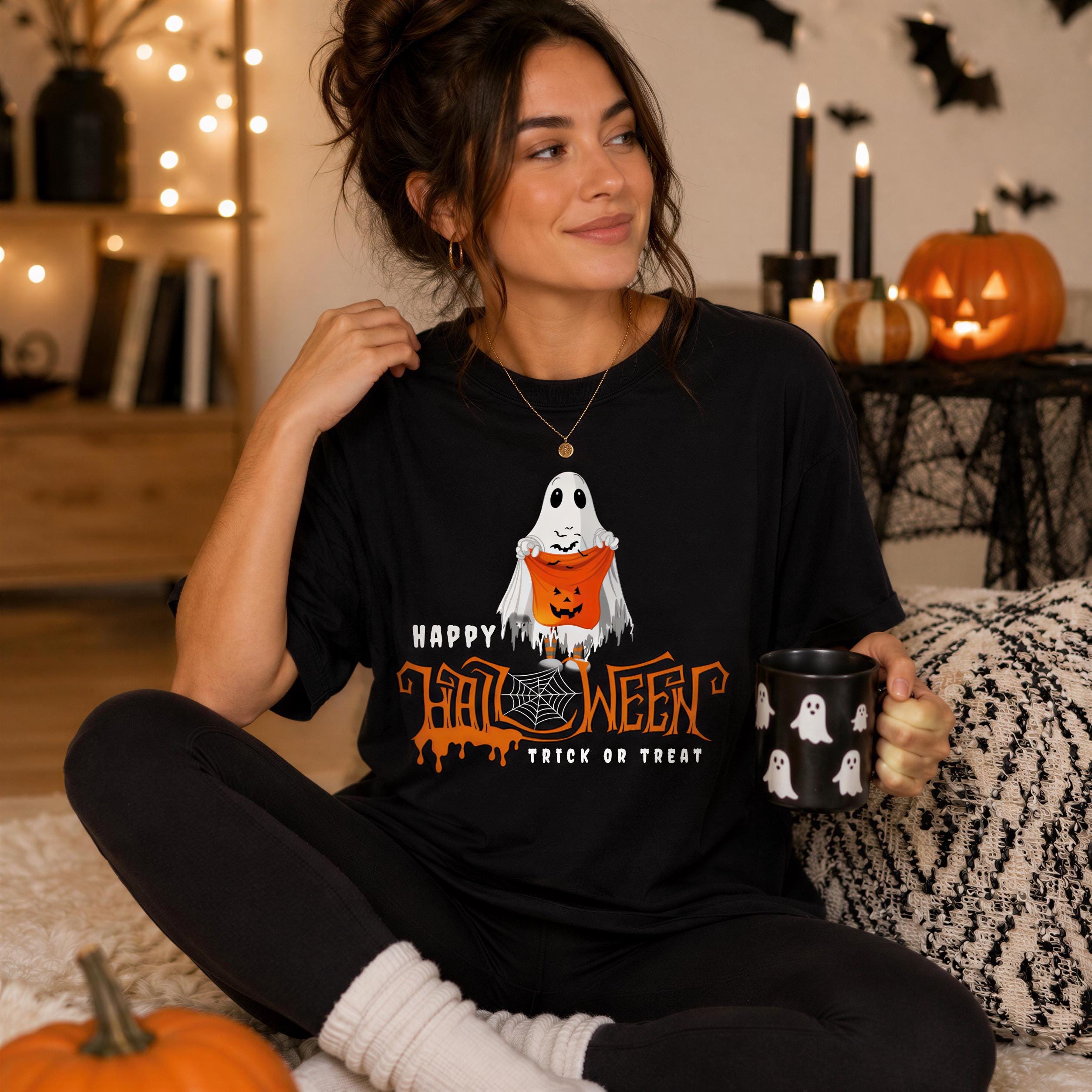 A woman sitting on a bed, wearing a black t-shirt with a Halloween-themed graphic and holding a mug.