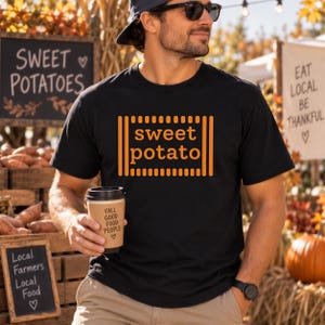 以下が含まれることがあります： 胸の中央にオレンジ色の「sweet potato」という文字が配置され、縦線と横線で囲まれた黒い半袖Tシャツ。Tシャツはベージュのパンツと黒い腕時計と合わせて着用されており、茶色の紙コップを手に持っている。