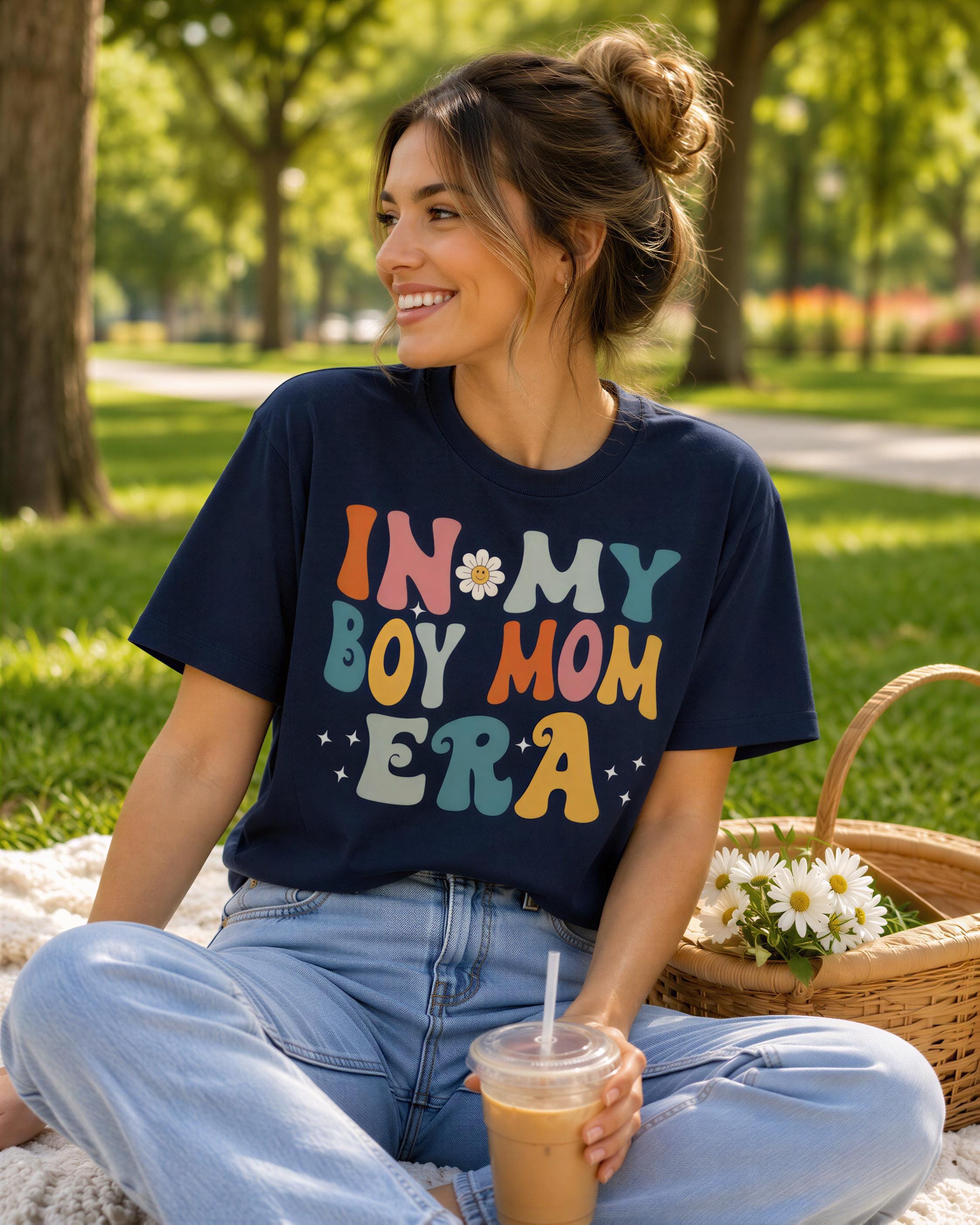 A woman sitting on a blanket in a park, wearing a blue t-shirt with the text "IN MY BOY MOM ERA" and holding a drink.