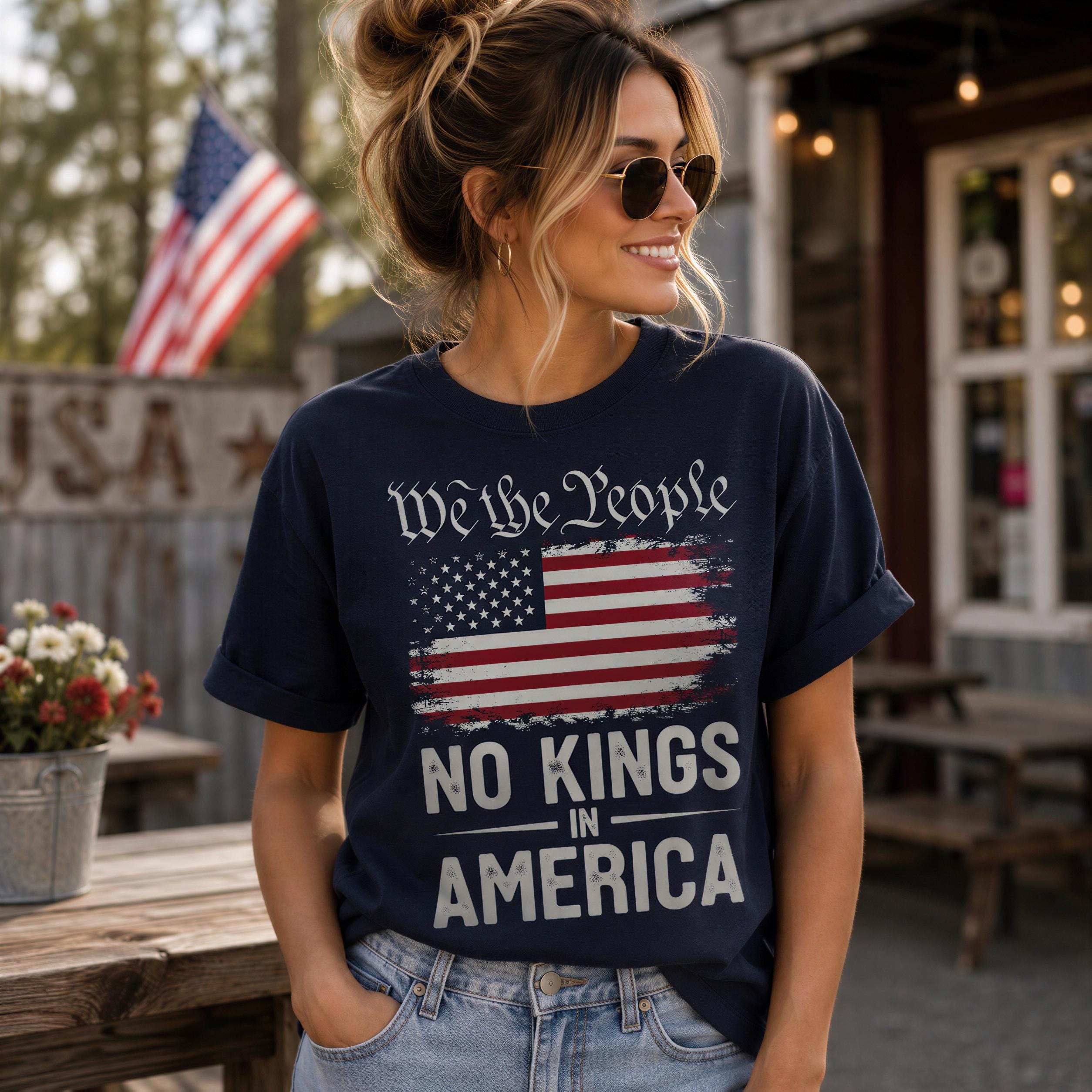 A woman wearing a navy blue t-shirt with the text "We the People" and "No Kings in America" printed on it, standing in front of a wooden fence with an American flag.