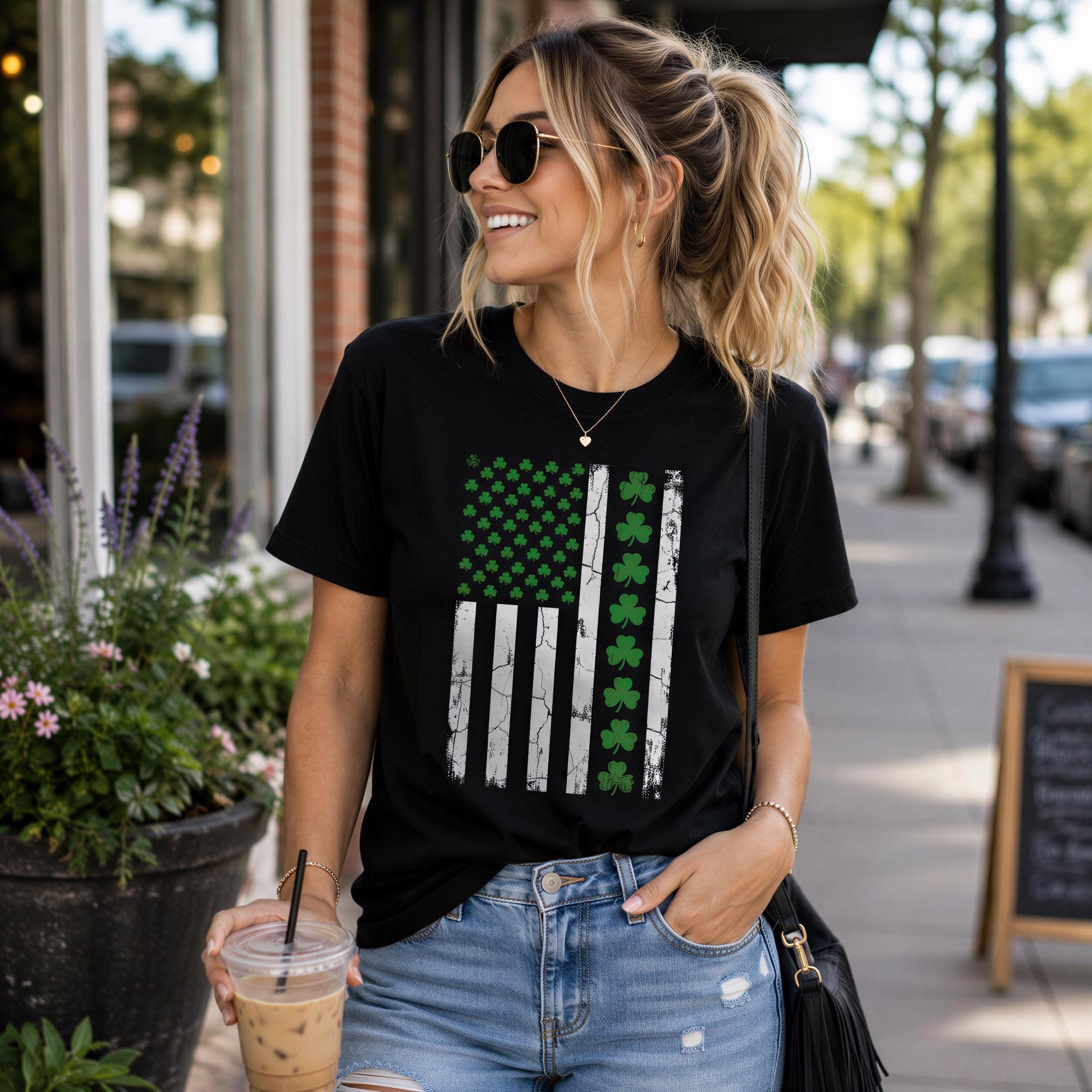 A woman wearing a black t-shirt with a green and white design, sunglasses, and holding a drink is standing on a city street.
