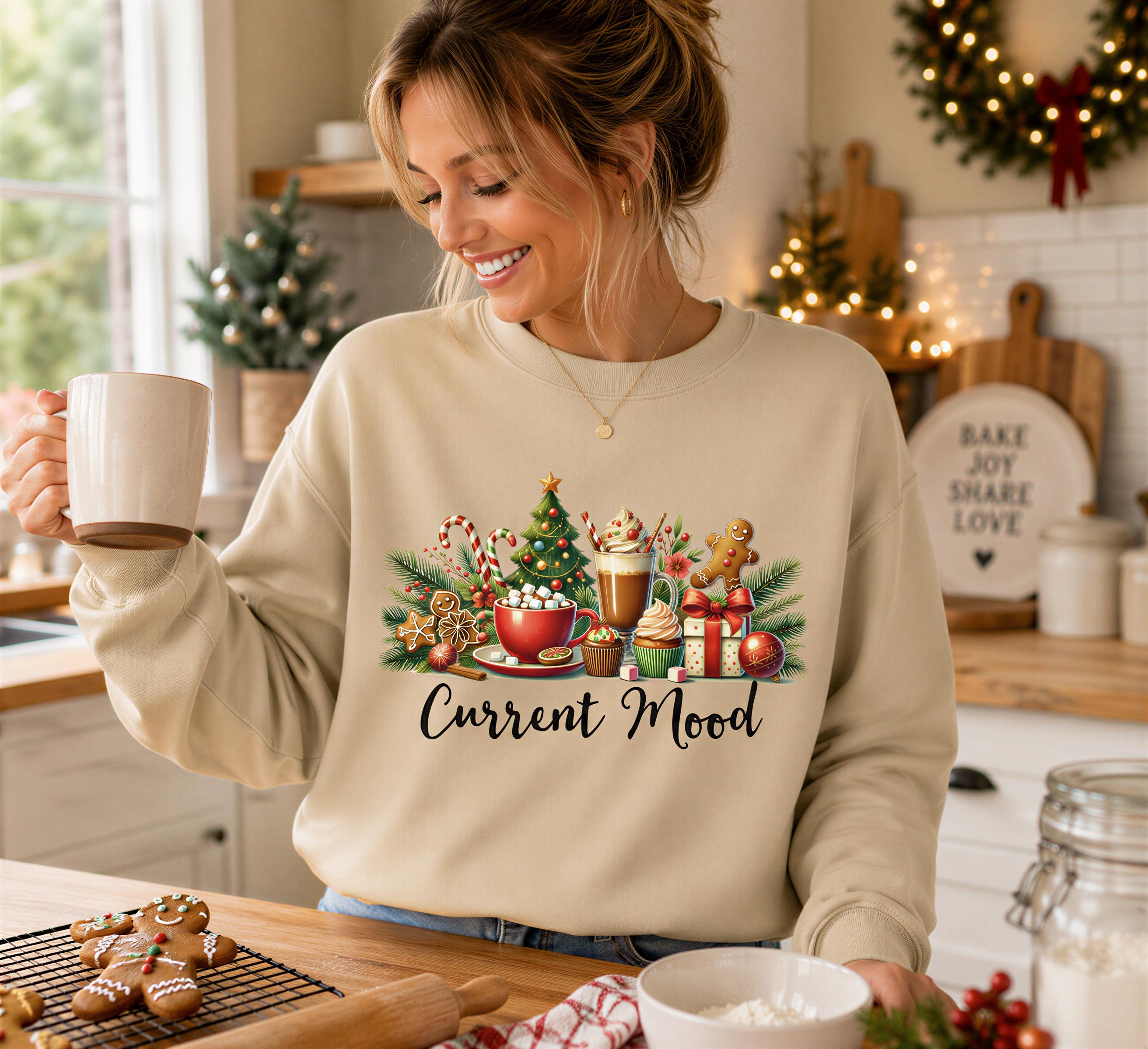 A woman wearing a beige sweater with a graphic design of Christmas-themed items, including a Christmas tree, gingerbread cookies, and a mug, is smiling and holding a mug in a kitchen setting.