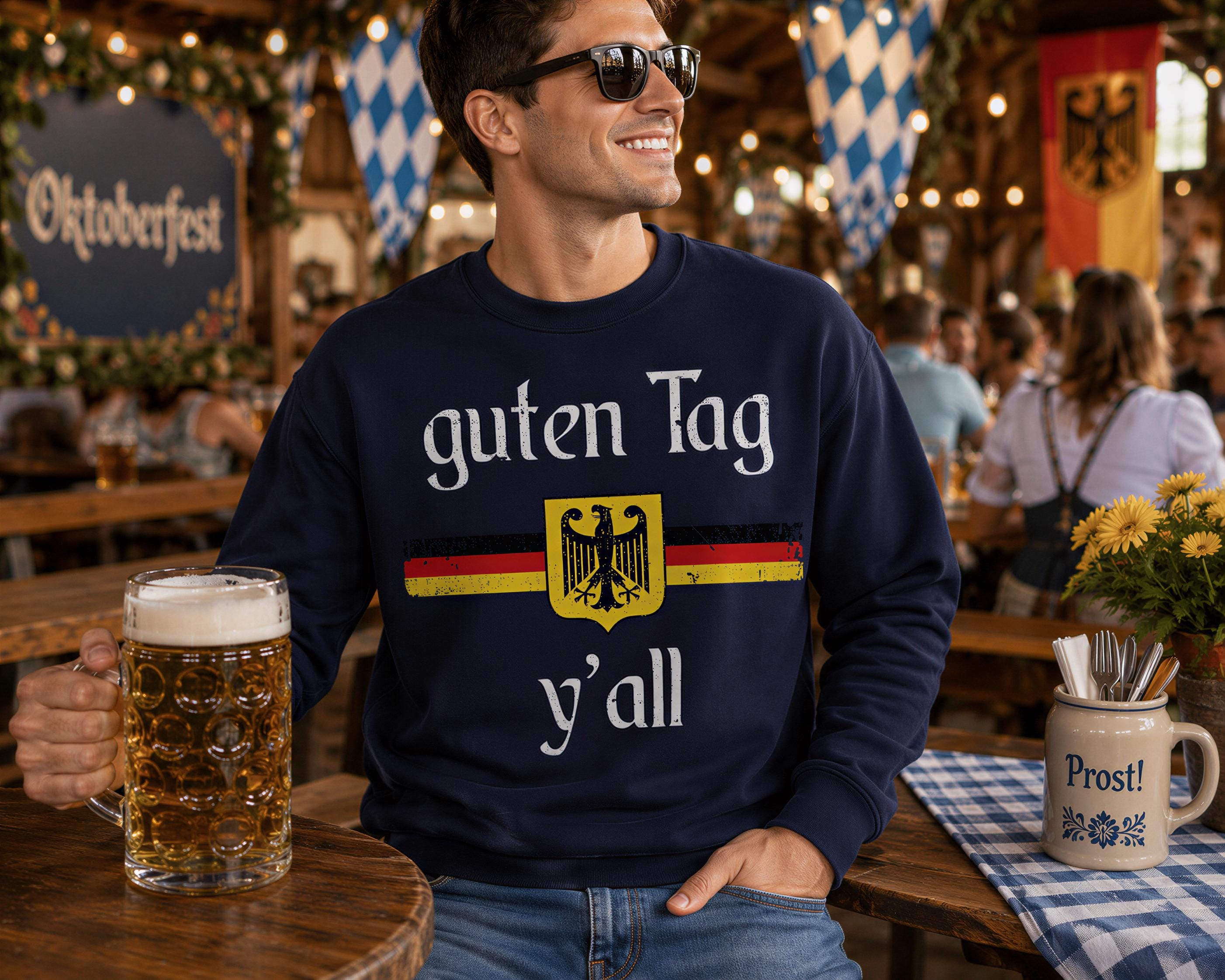 A man wearing a navy blue sweatshirt with a German flag design and the text "guten Tag y'all" is sitting at a table with a mug of beer in front of him.