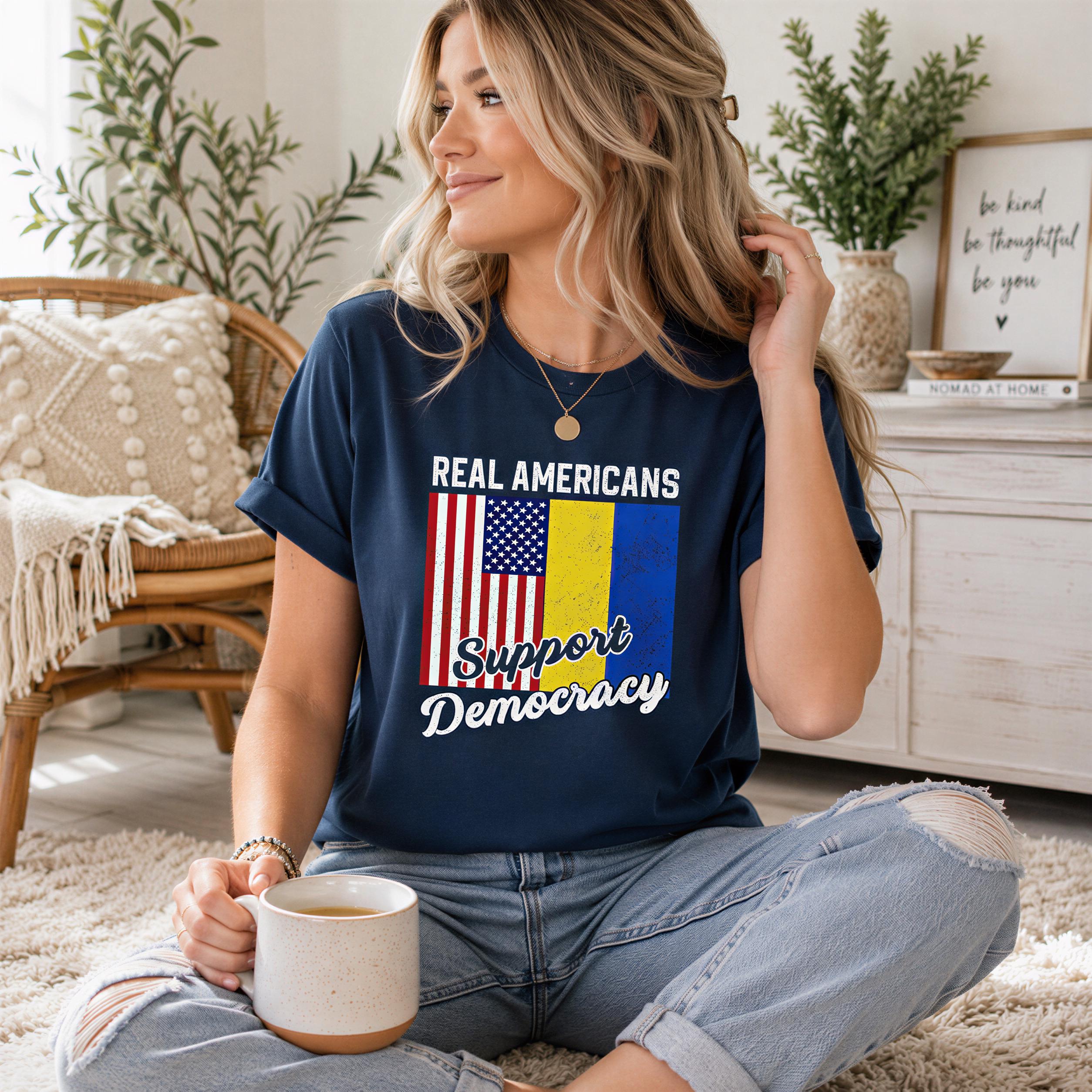 A woman wearing a blue t-shirt with the text "Real Americans Support Democracy" is sitting on the floor, holding a coffee mug and looking to the side.