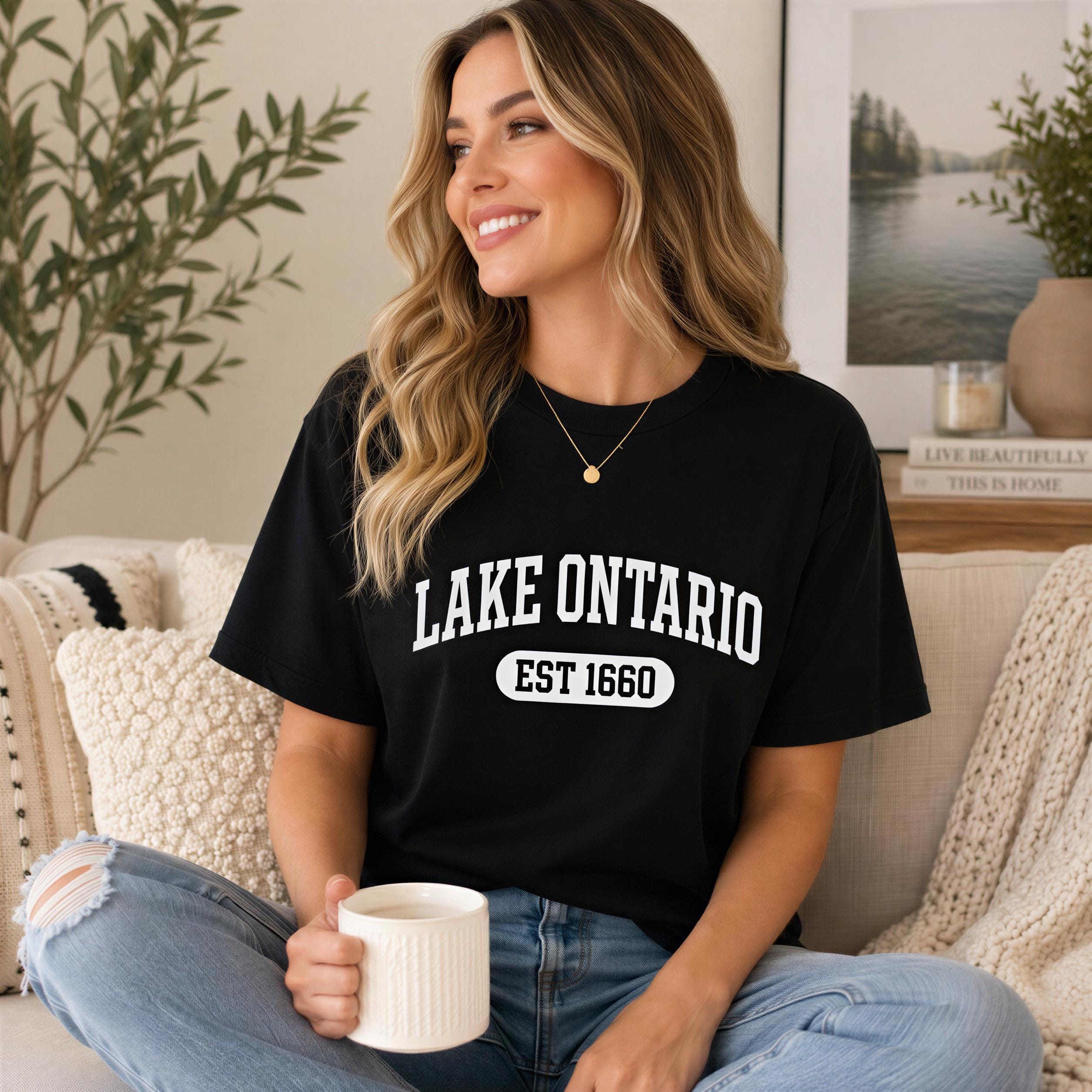 A woman wearing a black t-shirt with the text "LAKET Ontario EST 1660" is sitting on a couch, holding a white mug and smiling.