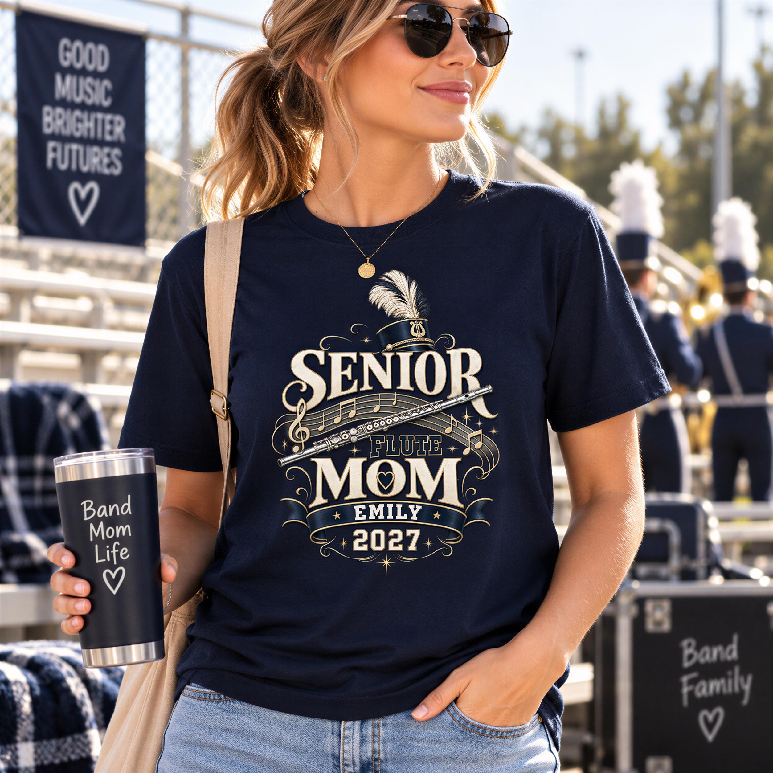 A woman wearing sunglasses and a navy blue t-shirt with a graphic design stands in front of a marching band, holding a cup with the text "Band Mom Life" on it.