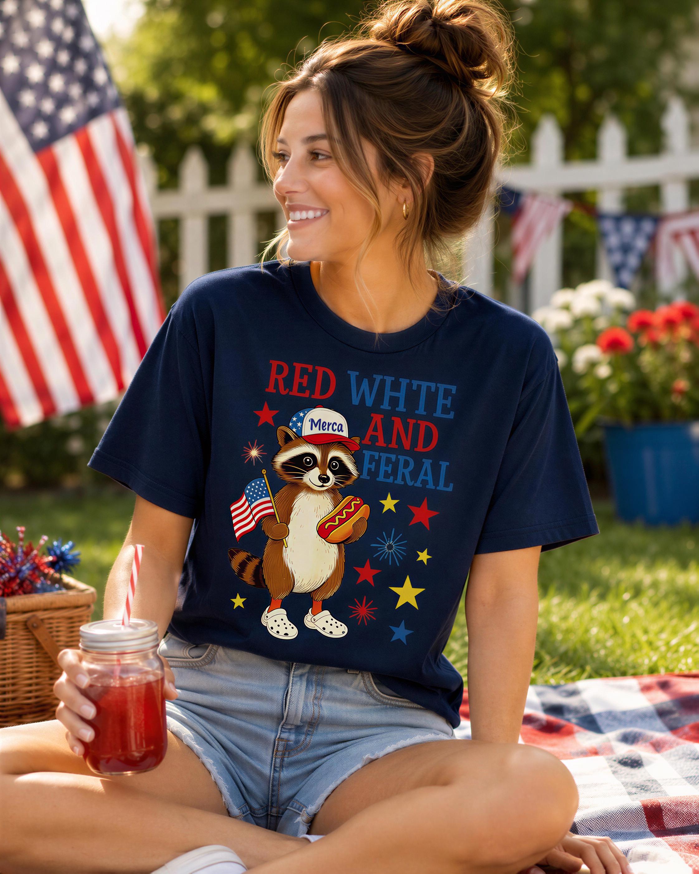 A woman wearing a blue t-shirt with a raccoon graphic and text, sitting on a blanket in a grassy area with an American flag in the background.