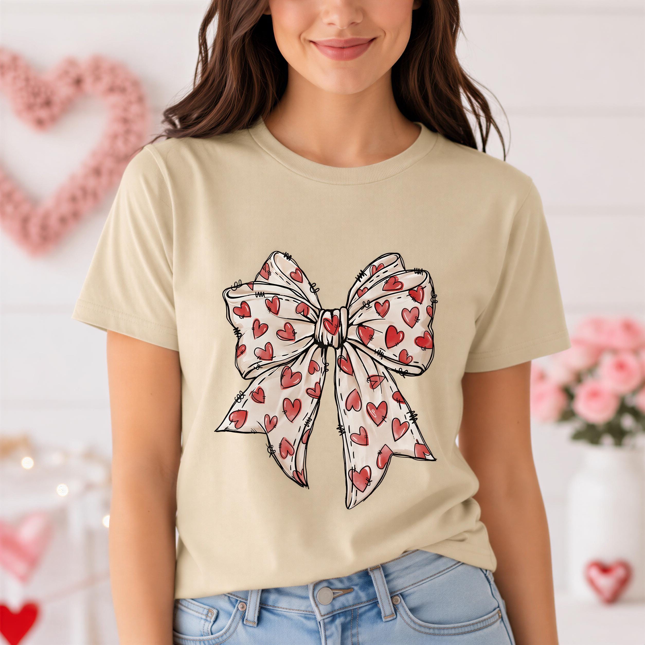 A woman wearing a beige t-shirt with a bow design featuring red hearts.