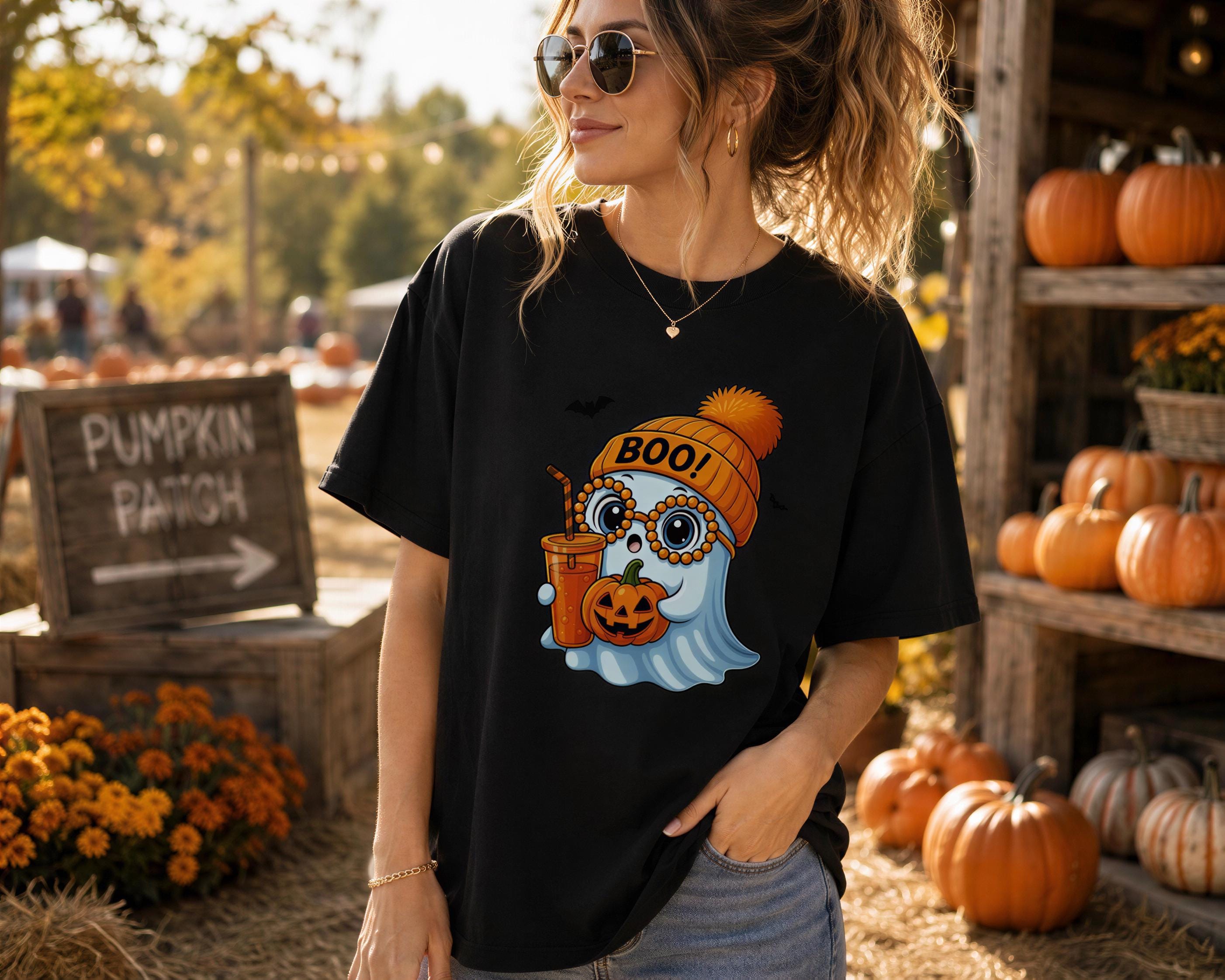 A woman wearing a black t-shirt with a graphic of a ghost wearing a hat and holding a pumpkin, standing in front of a pumpkin patch with various pumpkins and fall decorations.