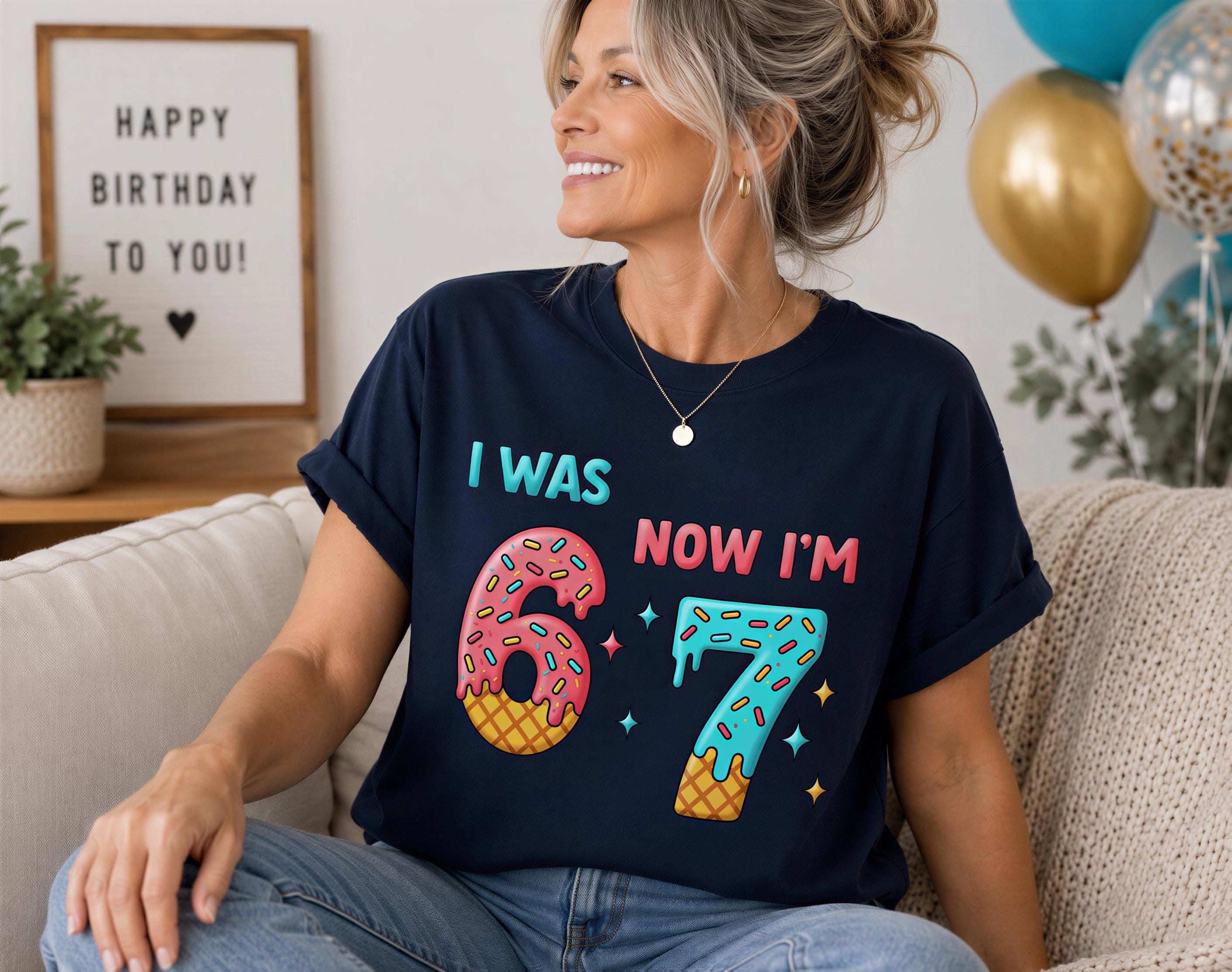 A woman sitting on a couch wearing a black t-shirt with a graphic design of a number 7 and the text "I WAS NOW I'M 67" printed on it.