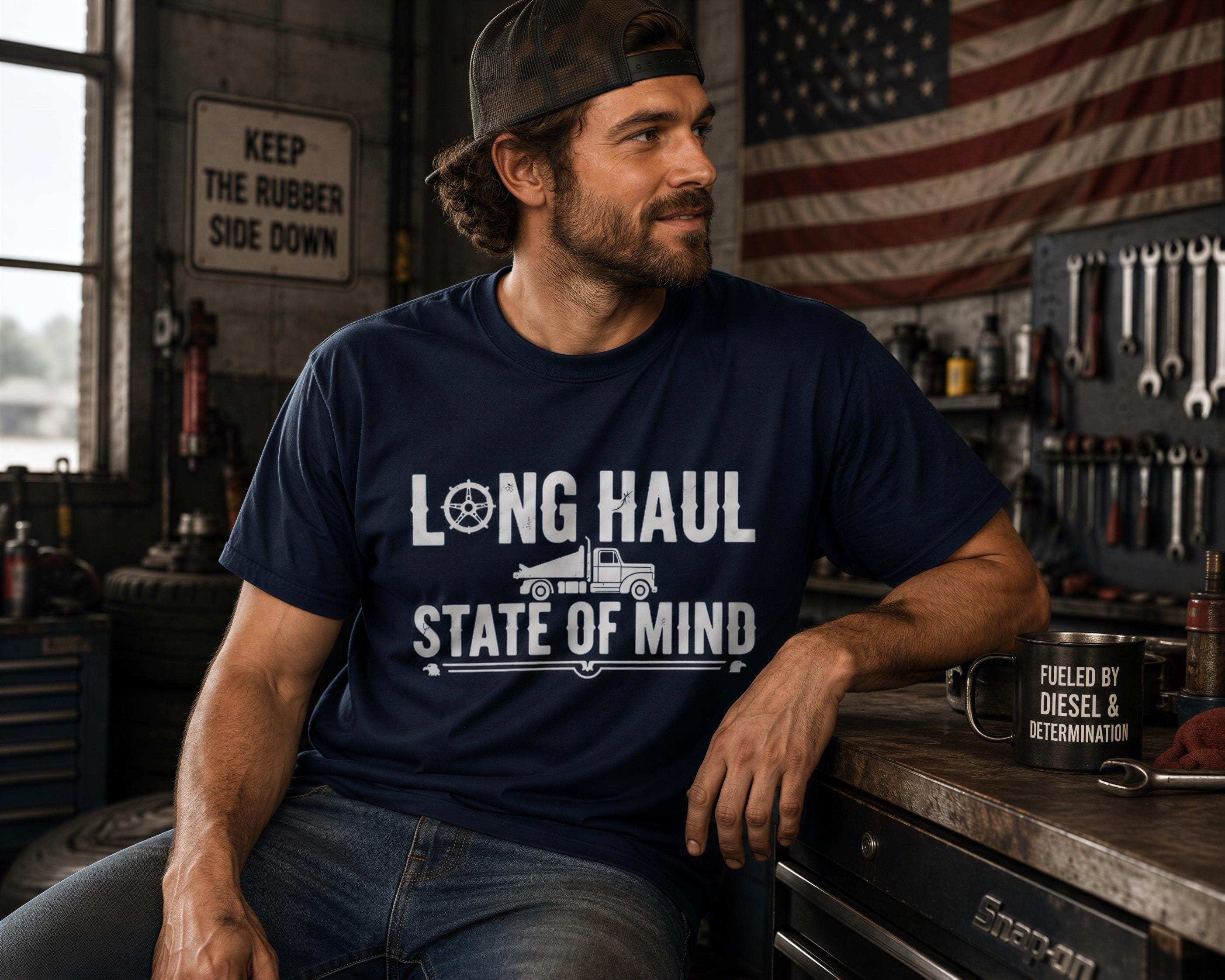A man wearing a blue t-shirt with the text "LONG HAUL STATE OF MIND" is sitting in a garage surrounded by tools and equipment.