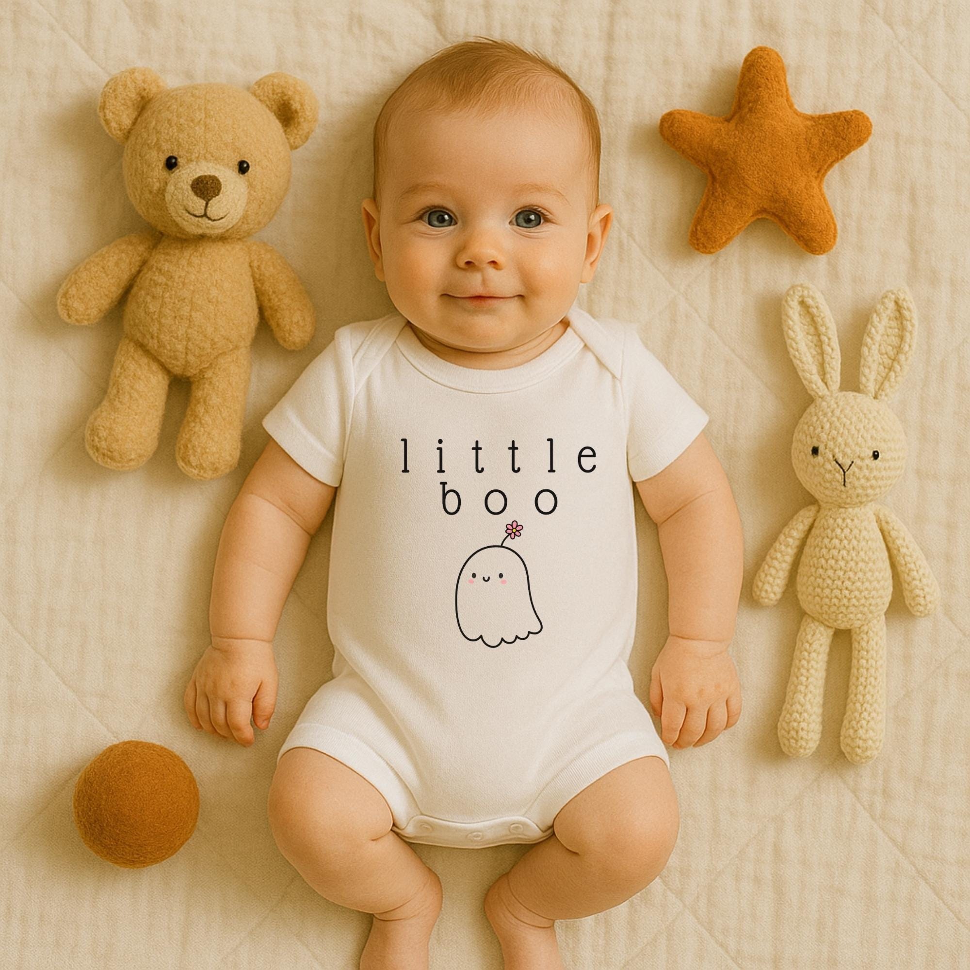 A baby wearing a white onesie with a ghost design lies on a bed surrounded by stuffed animals, including a teddy bear and a bunny.