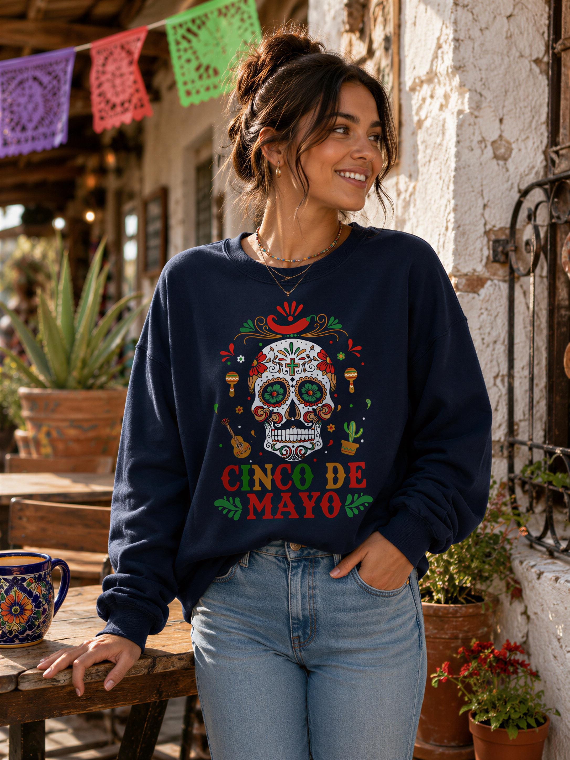 A woman wearing a navy blue sweatshirt with a colorful design of a skull and the text "CINCE DE MAYO" stands in front of a building with a festive decoration.