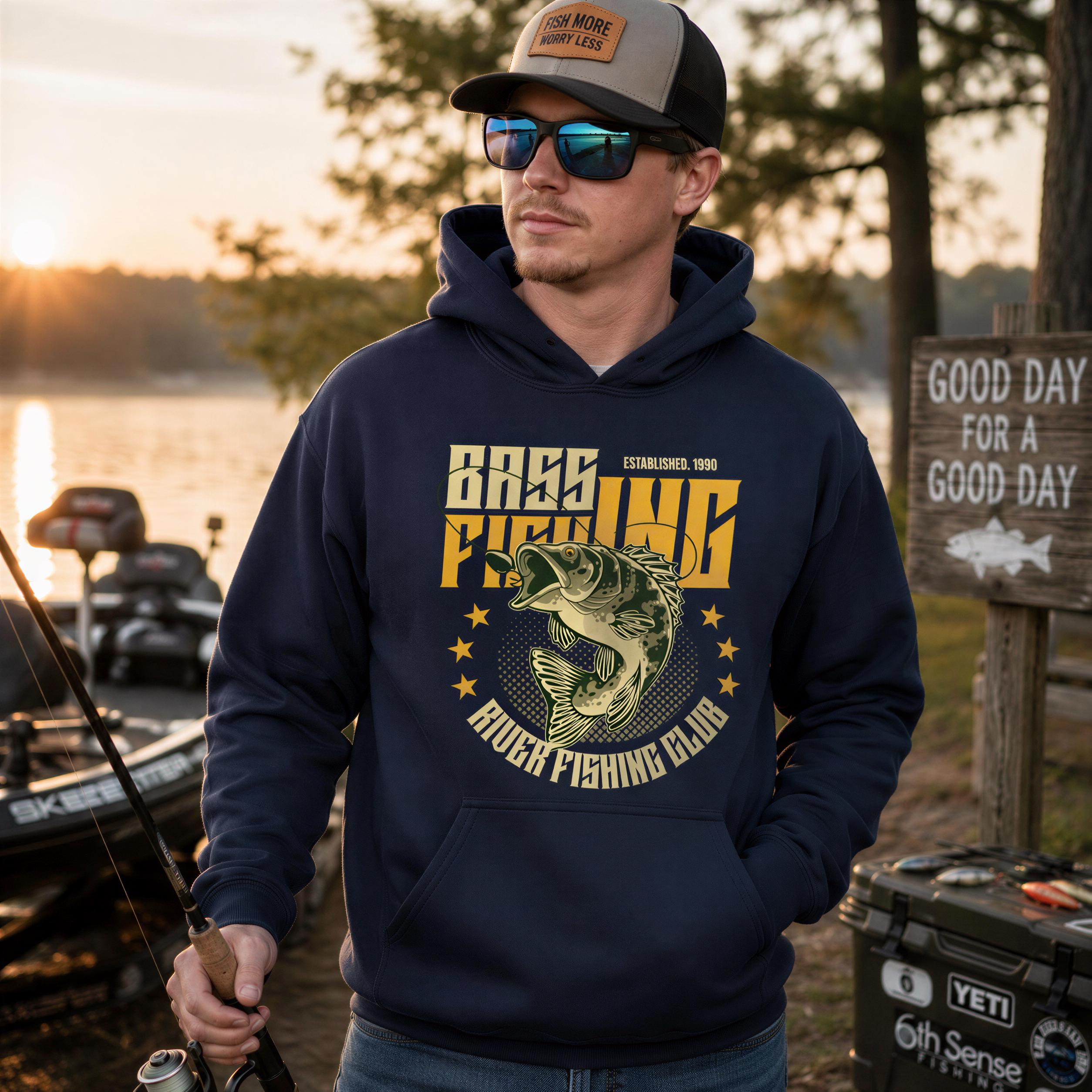 A man wearing a navy blue hoodie with a fishing-themed design stands near a lake, holding a fishing rod.