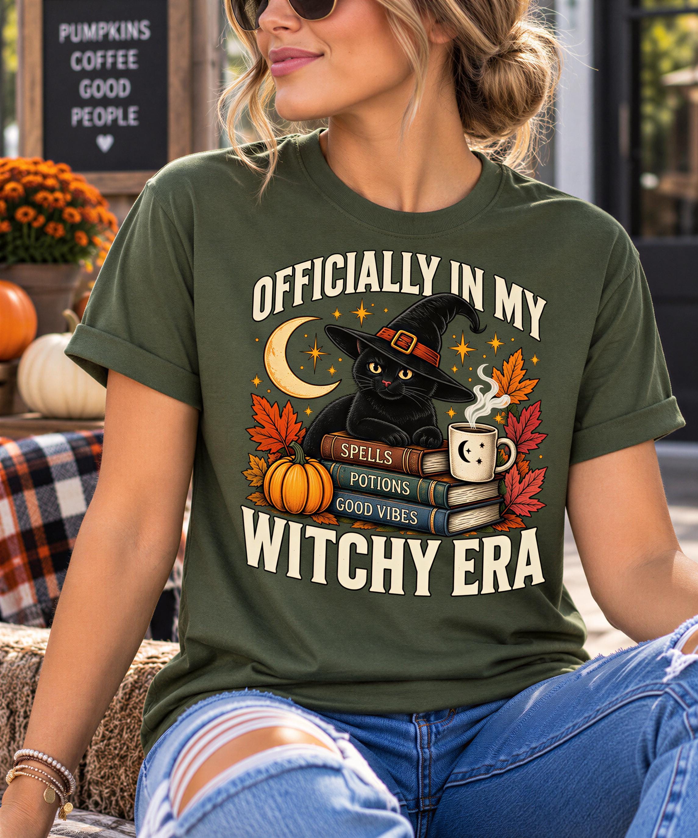 A woman wearing a green t-shirt with a Halloween-themed graphic and text, sitting on a bench with a pumpkin and other fall decorations in the background.