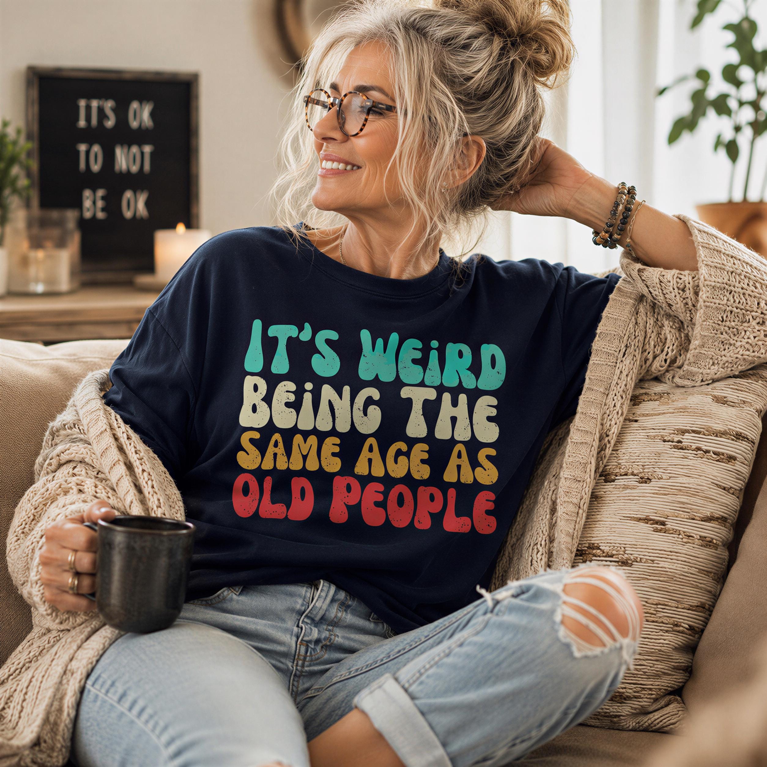 A woman sitting on a couch wearing a black t-shirt with a colorful text design, smiling and holding a black mug.