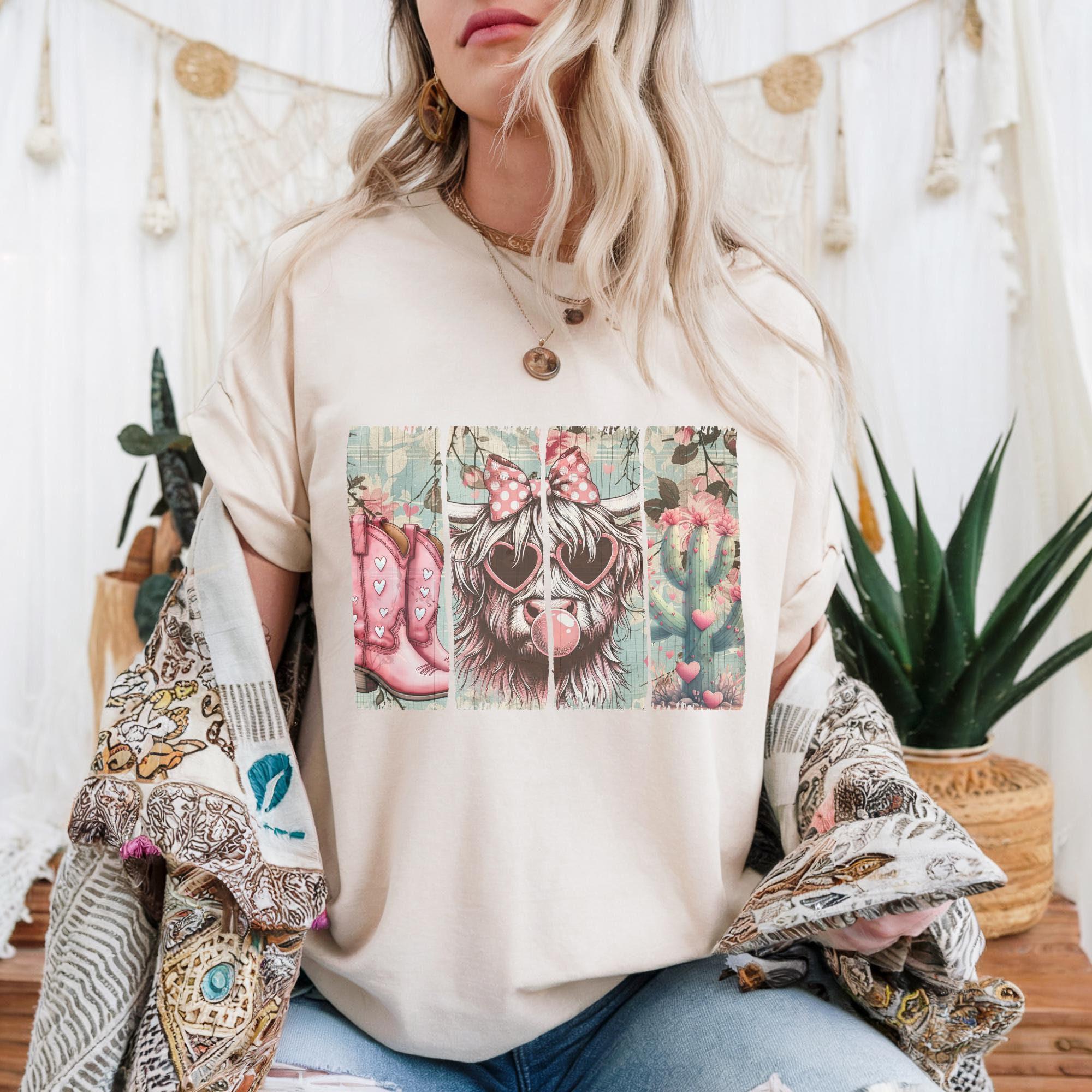 A woman wearing a white t-shirt with a graphic design of a dog wearing cowboy boots and sunglasses, sitting in front of a white curtain with plants and a dreamcatcher.