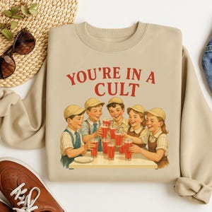A beige sweatshirt with a graphic design depicting a group of children happily drinking from glasses, with the text "YOU'RE IN A CULT" printed on it.