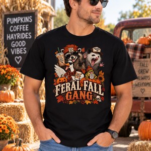 May include: A black t-shirt featuring a graphic of a raccoon, opossum, duck, and frog wearing autumn accessories and holding coffee cups. The text FERAL FALL GANG is printed below the animals, surrounded by pumpkins, sunflowers, and fall leaves.