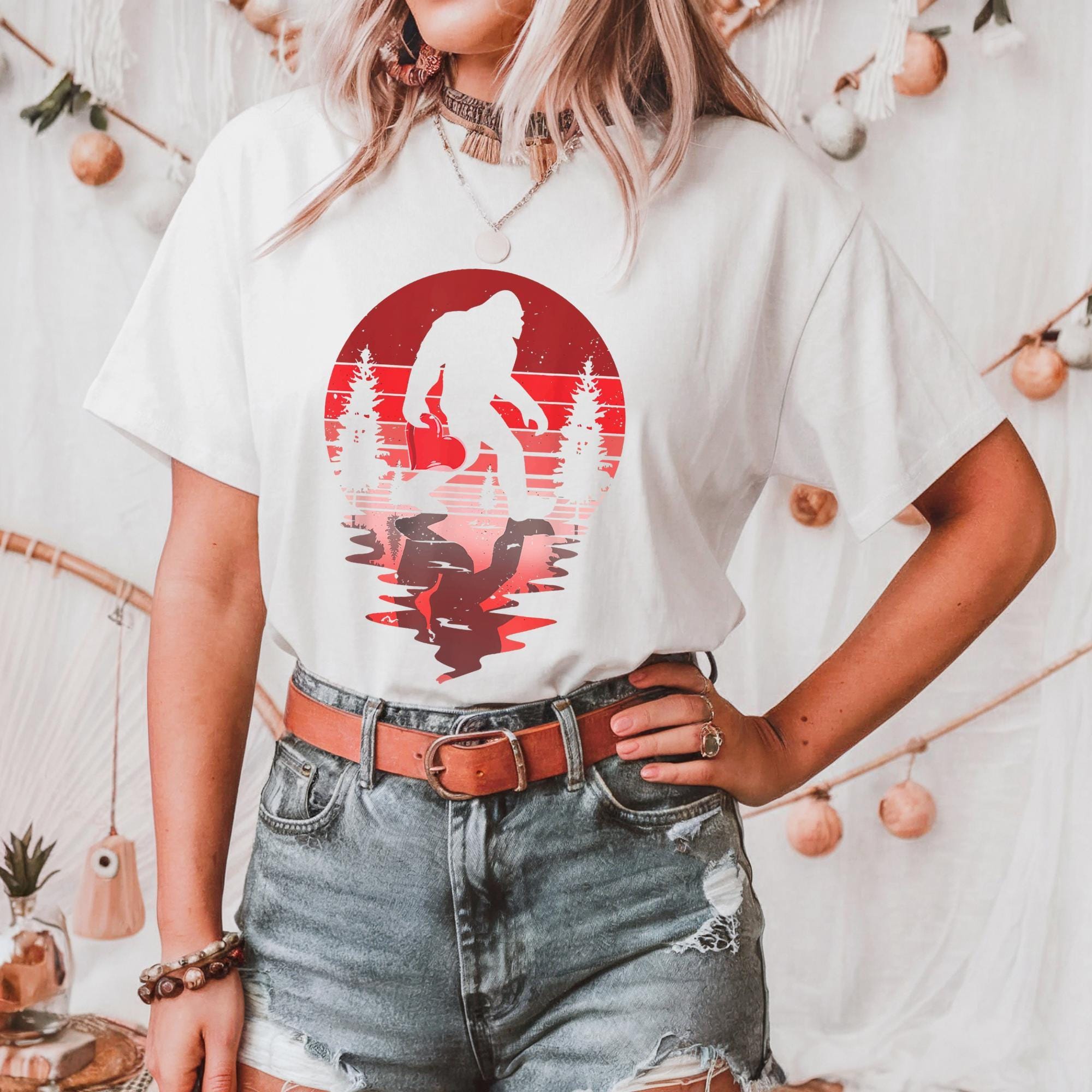 A woman wearing a white t-shirt with a graphic of a Sasquatch silhouette and a red and orange sunset design.