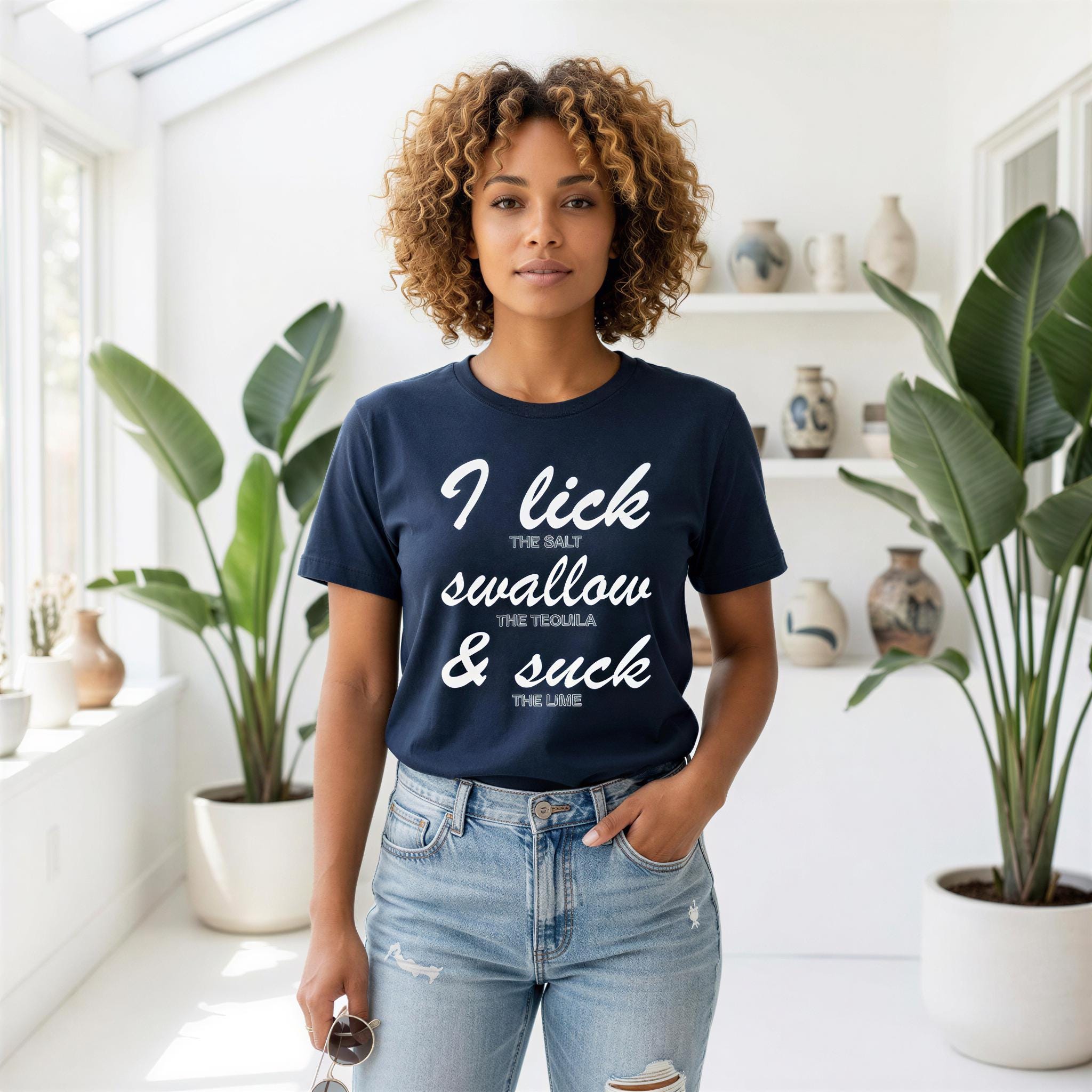 A woman with curly hair wearing a navy blue t-shirt with white text stands in a room with potted plants.