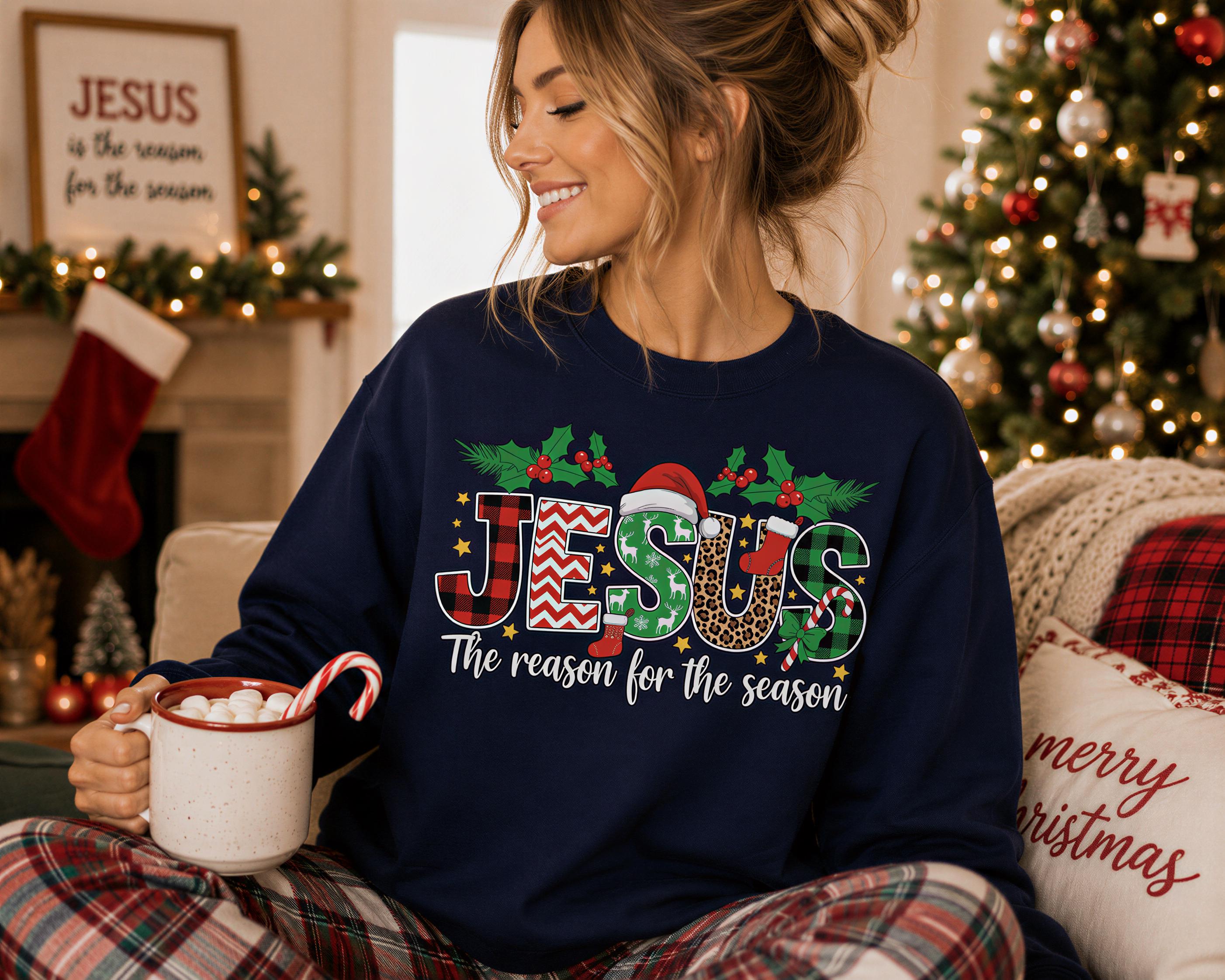 A woman wearing a navy blue sweatshirt with the text "JESUS The reason for the season" is sitting on a couch, holding a mug of hot chocolate and smiling.