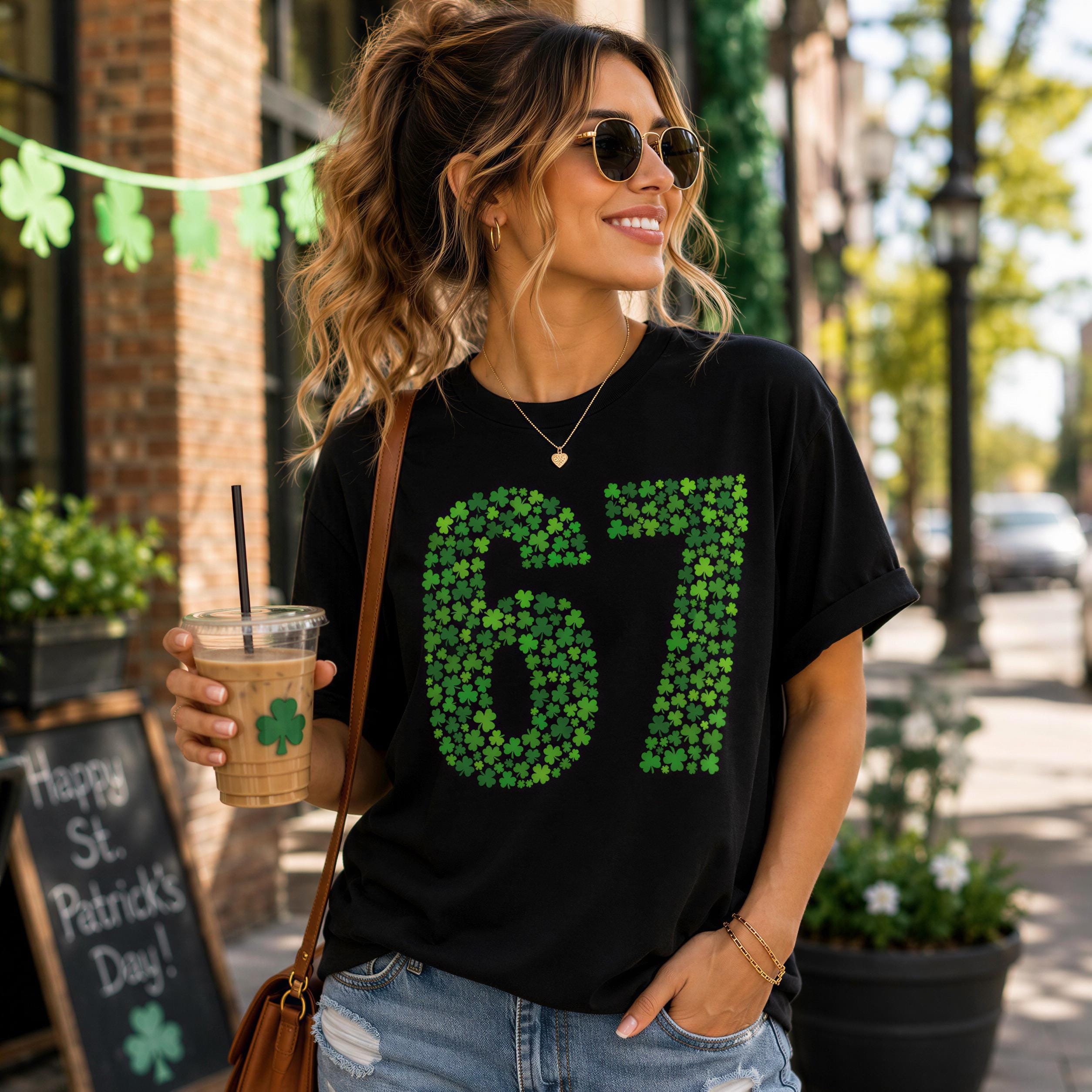 A woman wearing a black t-shirt with the number 67 and sunglasses is holding a drink and standing on a city street.