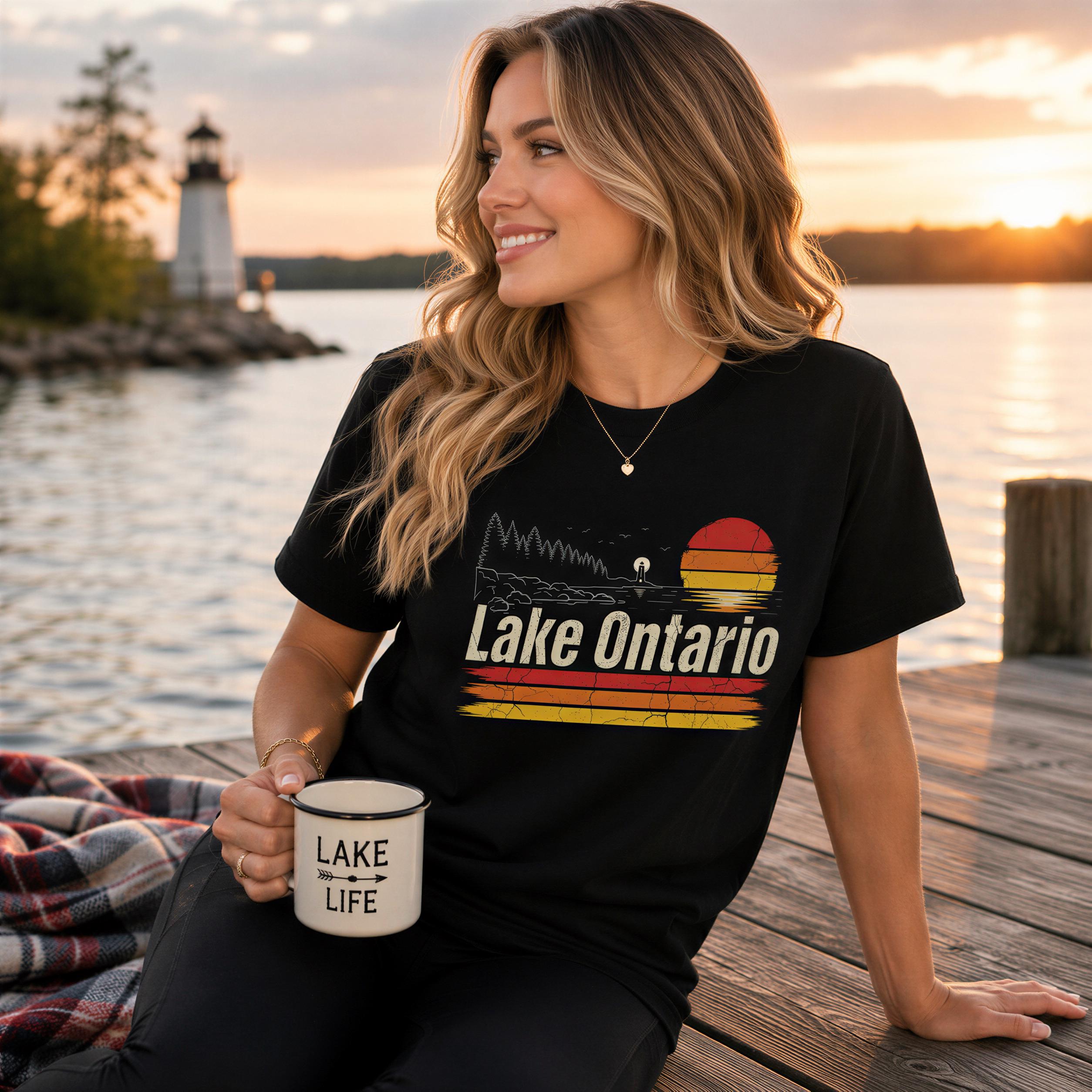 A woman is sitting on a dock, holding a mug and smiling at the camera. She is wearing a black t-shirt with a graphic design that says "Lake Ontario".