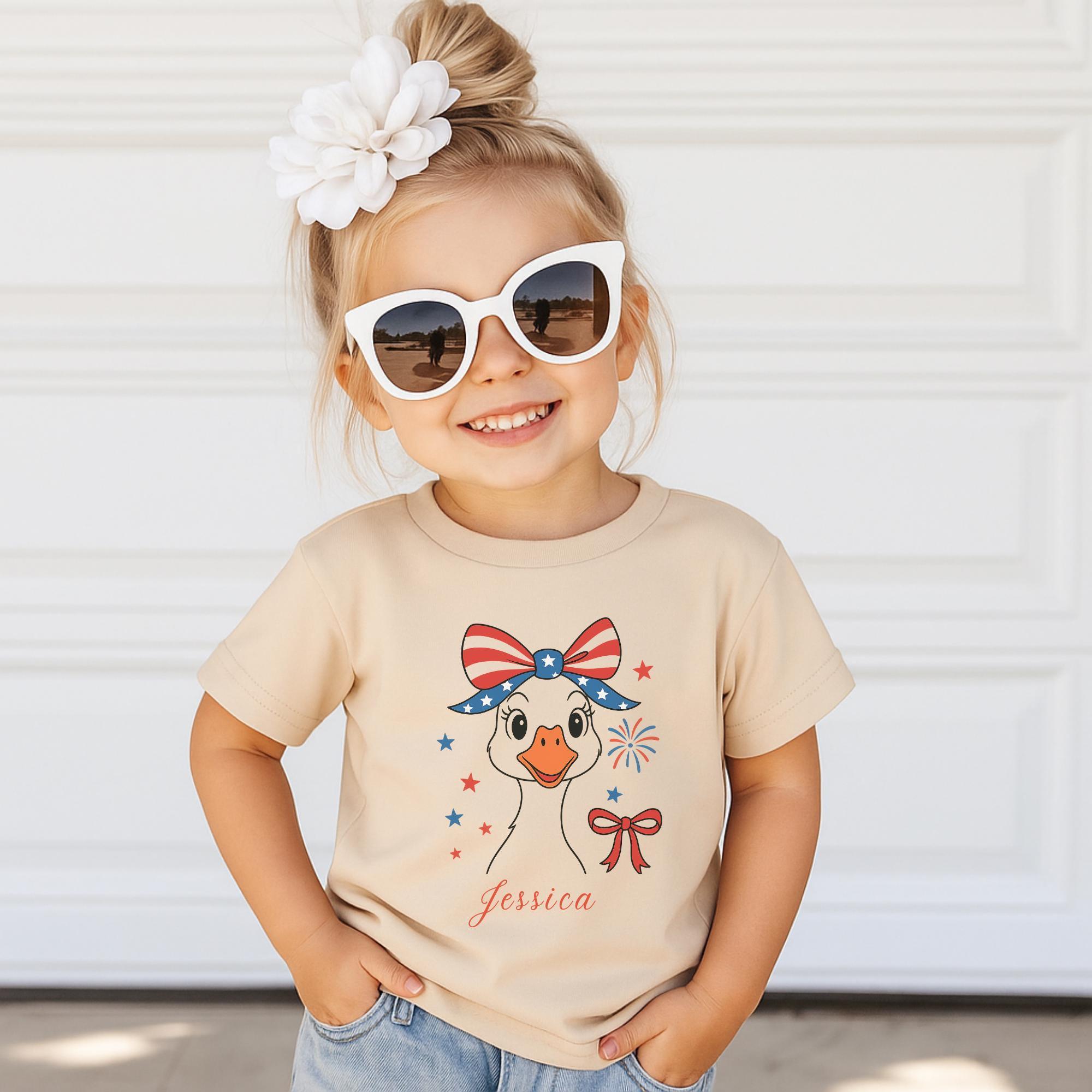 A young girl wearing a beige t-shirt with a cartoon duck and patriotic design, smiling and posing in front of a white garage door.