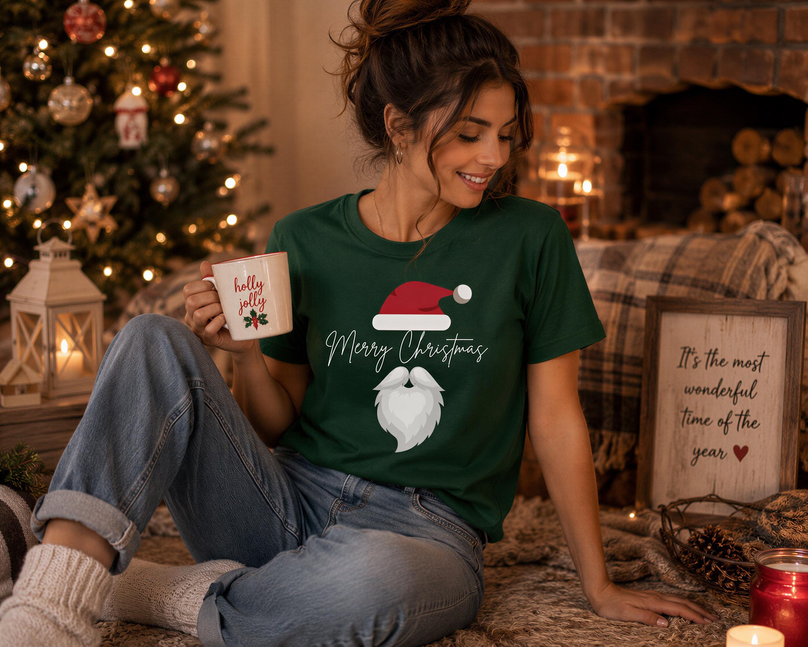 A woman in a green t-shirt with a Santa hat and the text "Merry Christmas" is sitting on the floor, holding a mug and smiling. The background includes a Christmas tree, a fireplace, and other holiday decorations.