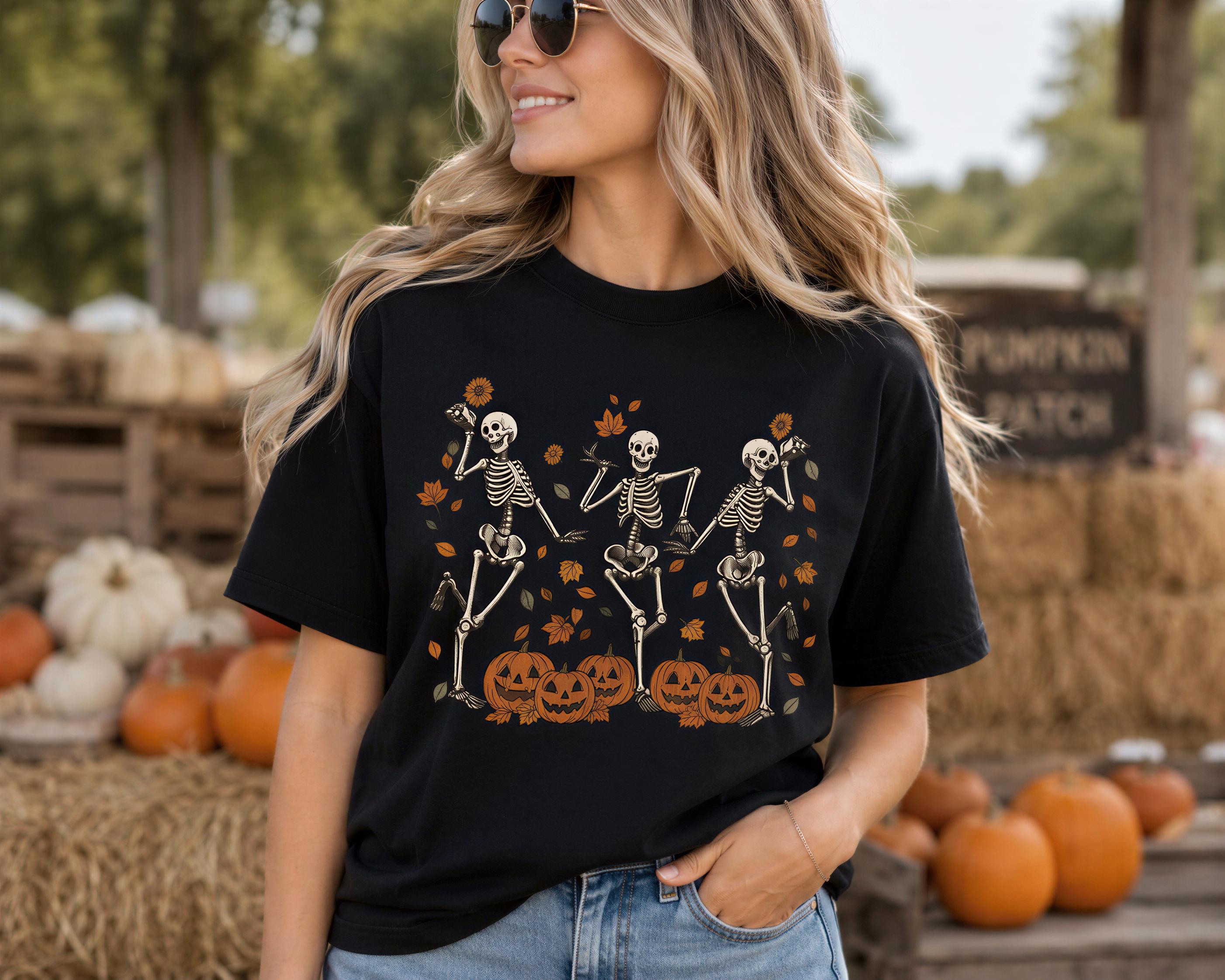 A woman wearing a black t-shirt with a Halloween-themed design of three skeletons dancing with pumpkins in a field.