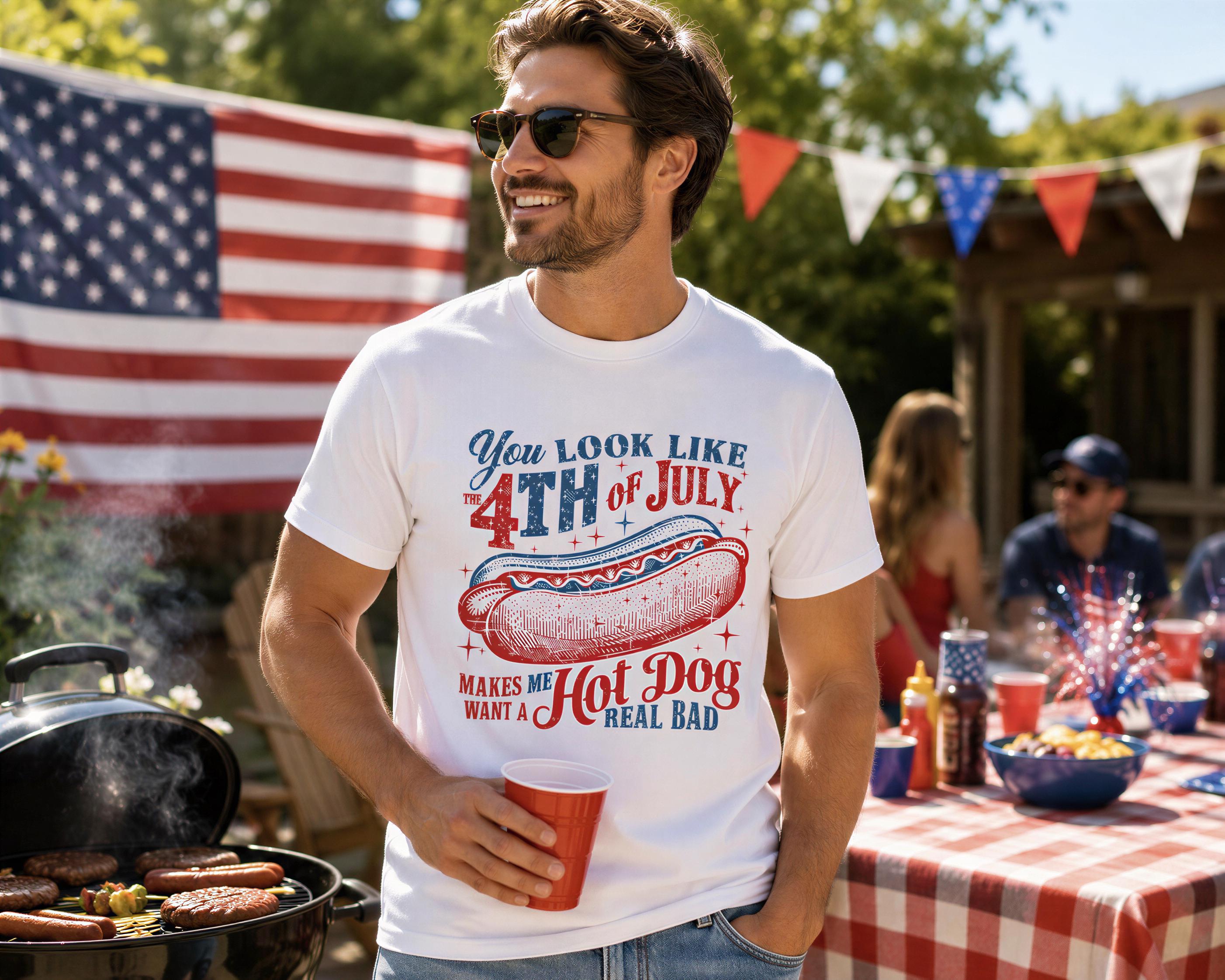 A man wearing a white t-shirt with a graphic of a hot dog and text, smiling and holding a red cup, standing in front of an American flag and a table with food and drinks.